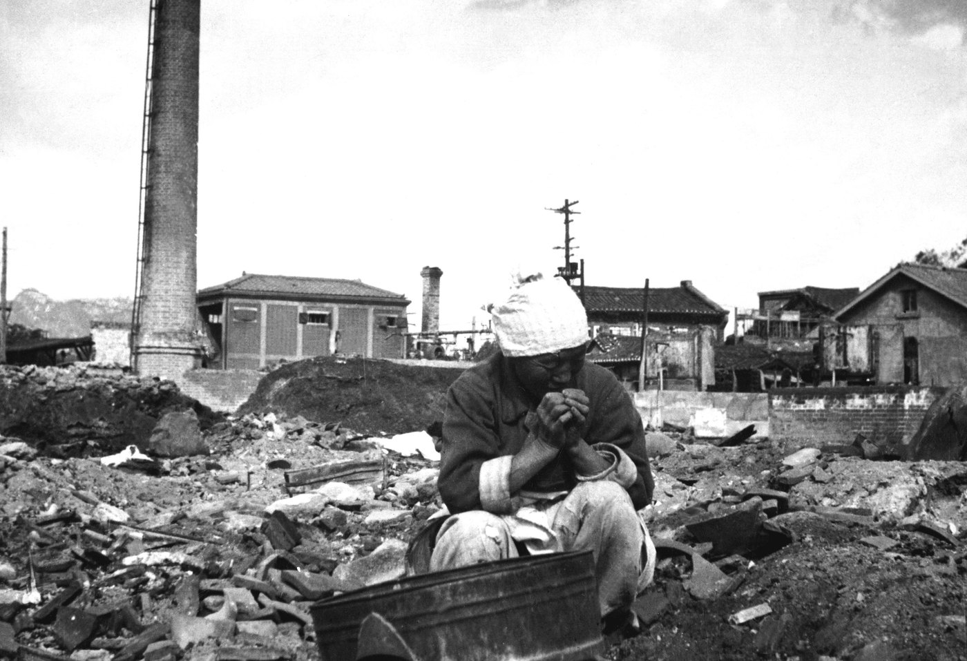 Black and white photograph of an elderly Korean woman in a head scarf crouching among rubble, with damaged brick buildings and a tall chimney behind her.