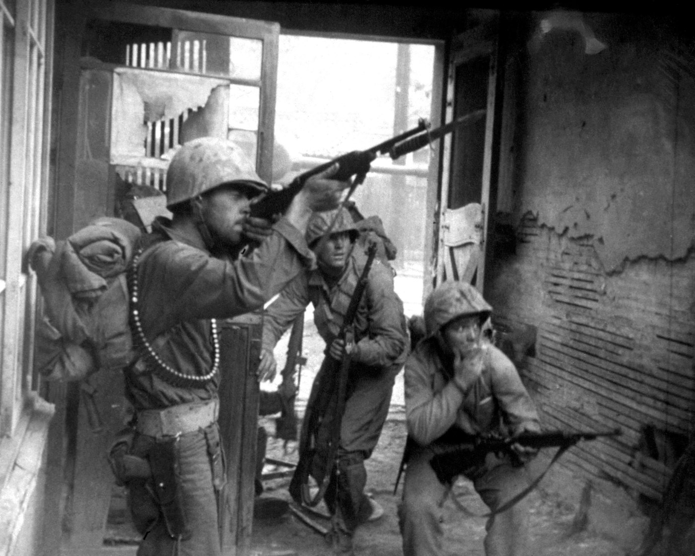 Black and white photograph of three helmeted soldiers in a ruined street; the one in front aims a rifle upward past a doorway while another crouches against a wall.