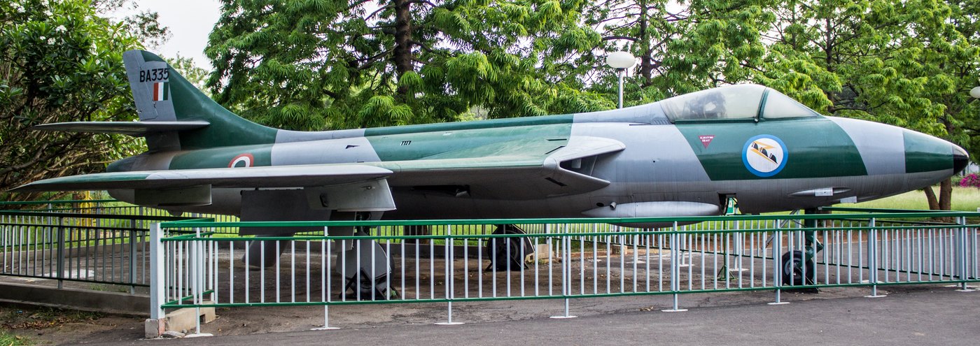 Green and gray Hawker Hunter jet with Indian Air Force roundels and the serial BA335 parked behind a green fence among trees