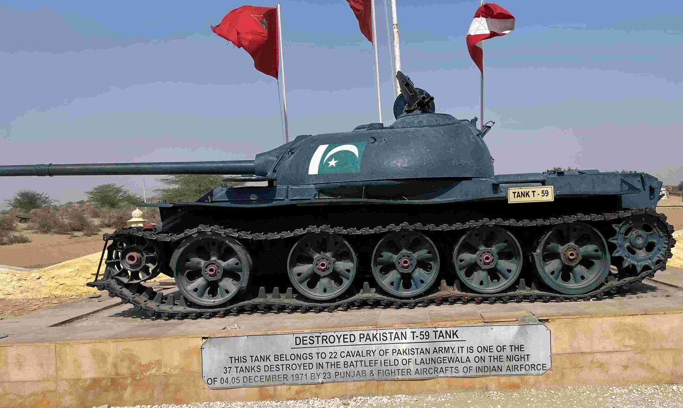 Dark green T-59 tank painted with a Pakistani flag standing on a stone plinth with a metal plaque, red flags flying behind it