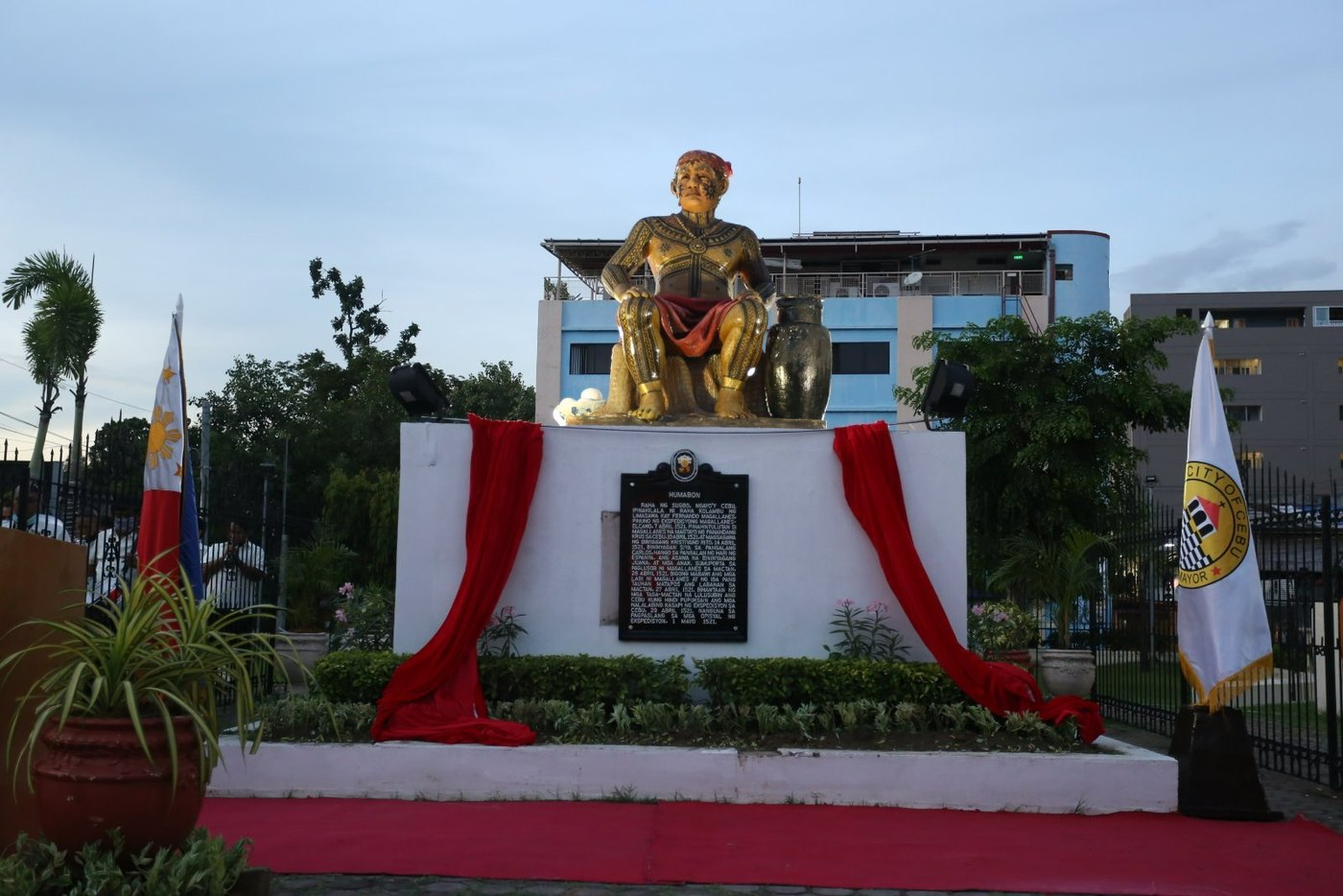 Gilded statue of a seated man in ornate armor beside a large jar, on a white pedestal with a black historical marker, draped in red cloth, with a Philippine flag and a Cebu City flag on either side