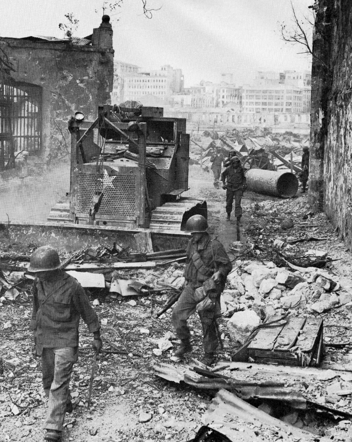 Helmeted American soldiers with rifles pick their way through rubble and twisted sheet metal beside a bulldozer marked with a white star, between a shattered stone wall and the gutted shells of tall buildings