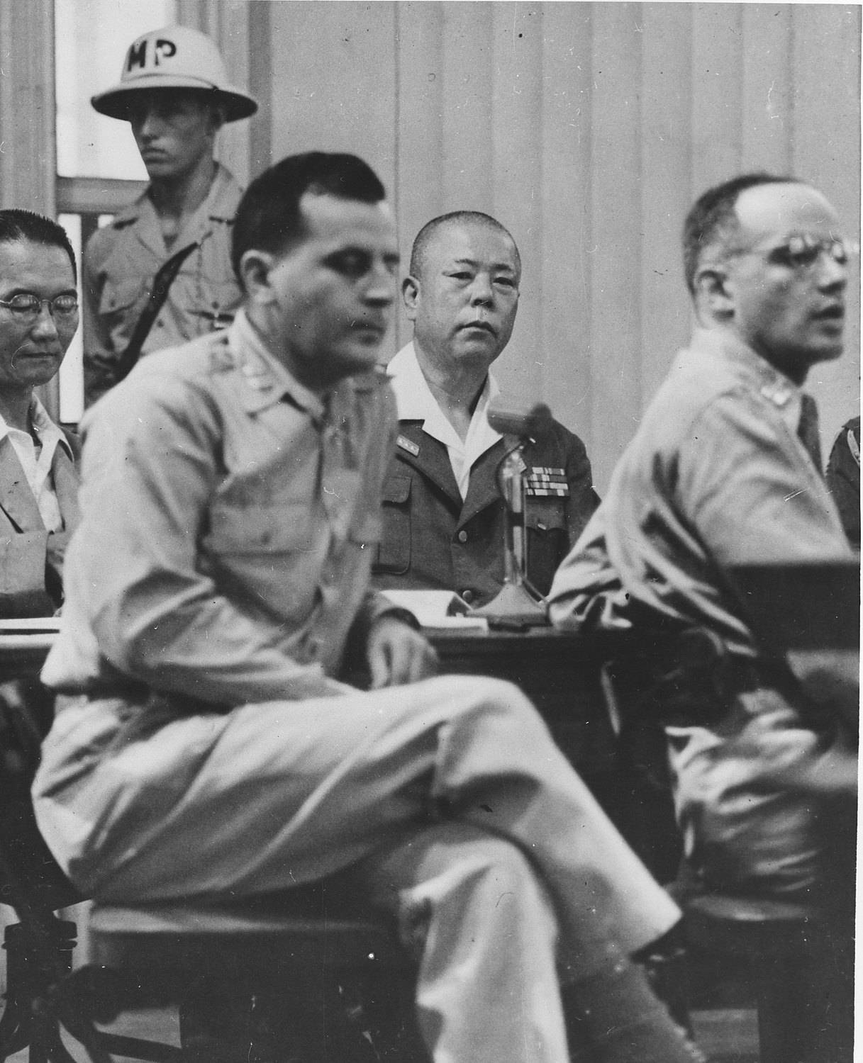 A Japanese general in uniform with rows of medal ribbons sits behind a microphone at a table, between American officers in khaki and an older man in glasses, while a military policeman in a white helmet marked MP stands behind him