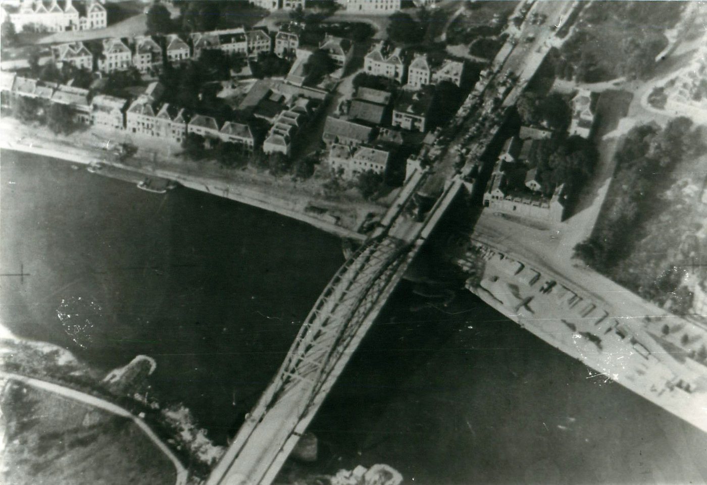 Black and white aerial photograph of a long road bridge across a river with burnt out vehicles at one end