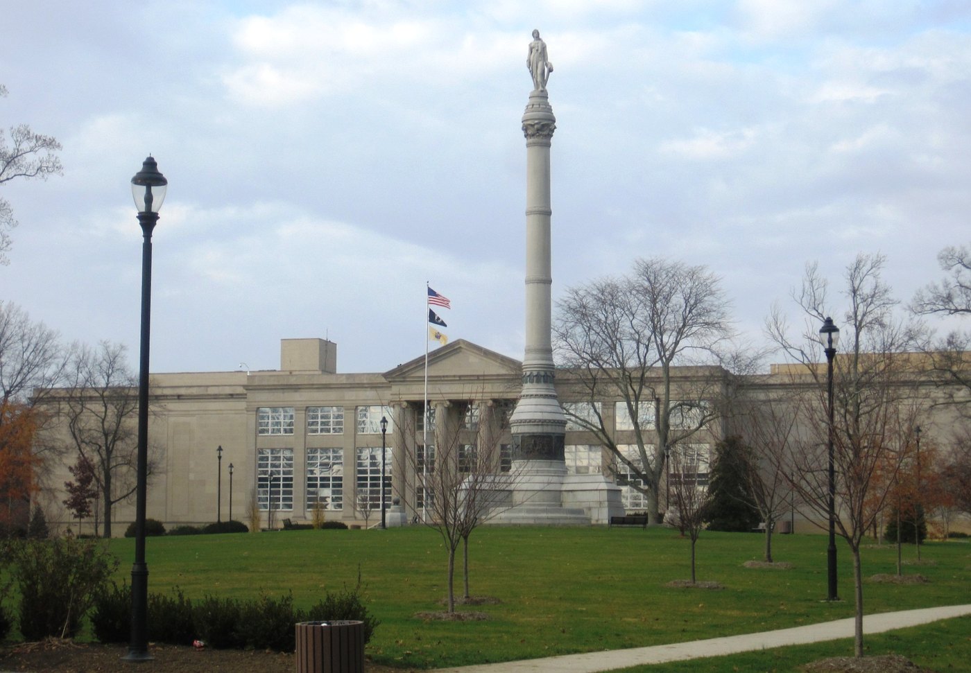 A tall granite column topped by a statue rises from a lawn in front of a courthouse with a flagpole and bare trees