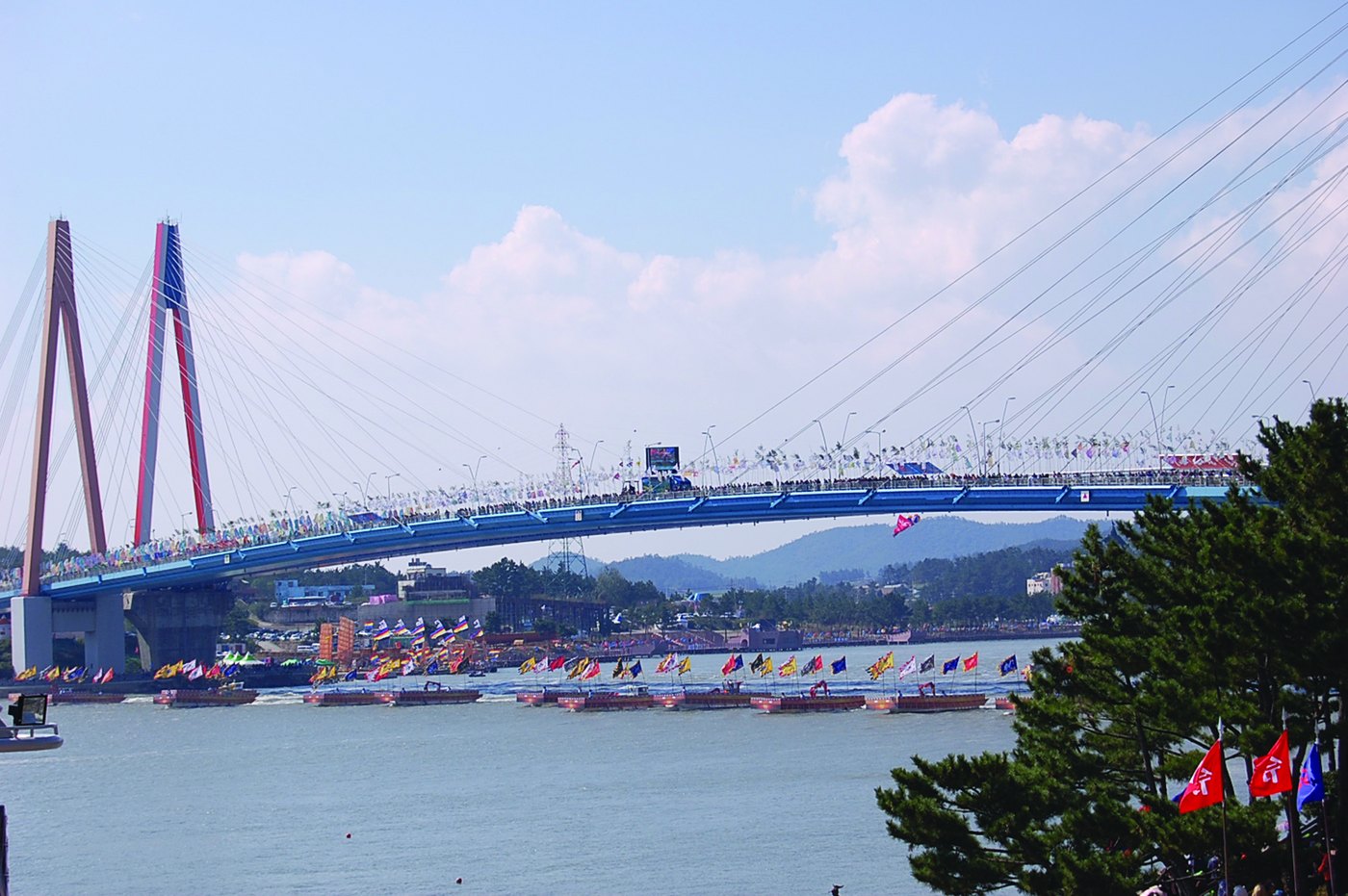 A long cable-stayed bridge with red and blue pylons crowded with people and flags, above a channel where a line of flat boats decked with colorful banners floats in front of a shore covered in flags