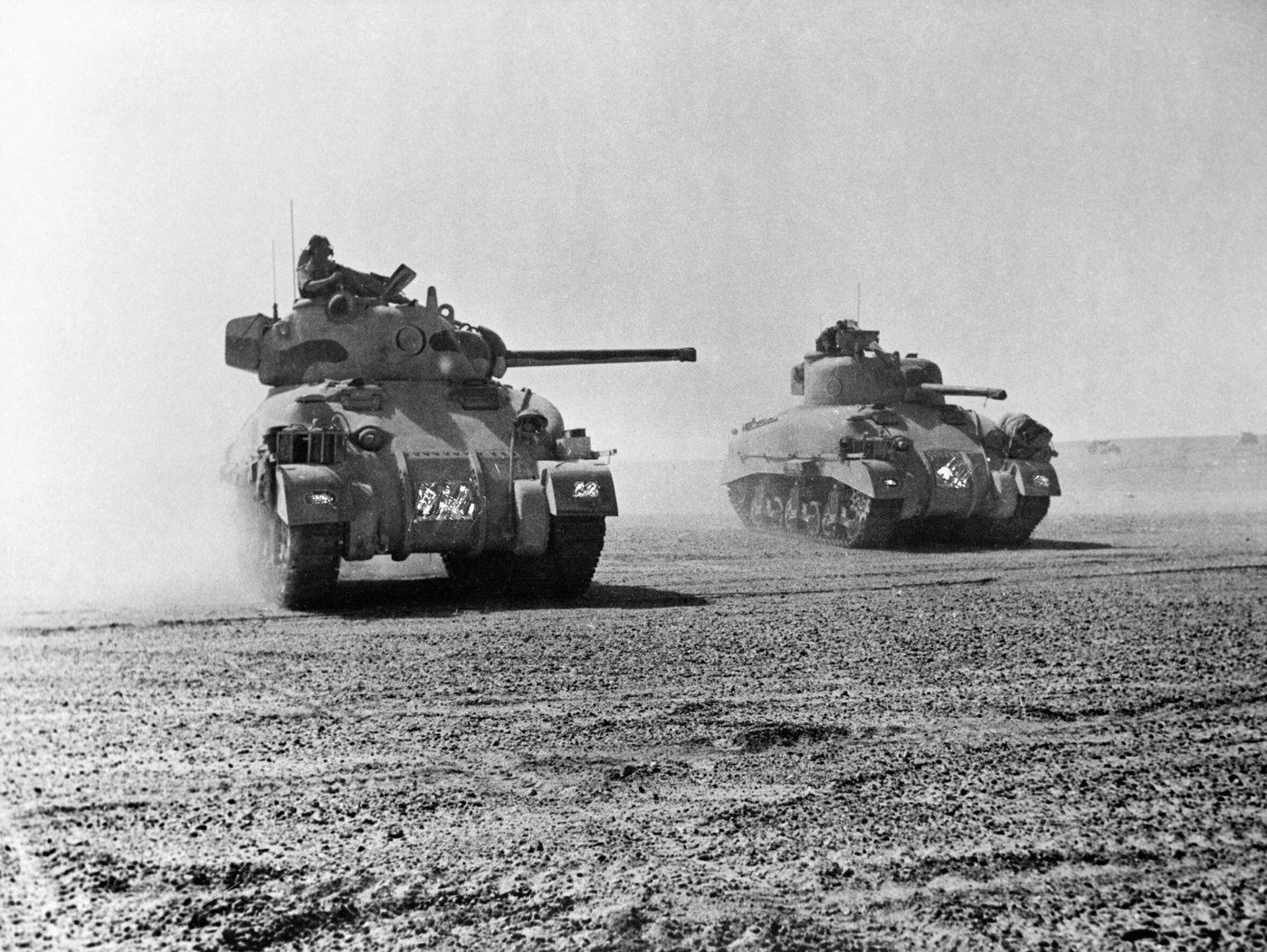 Black and white photograph: two Sherman tanks drive across flat, stony desert through clouds of dust, a crewman sitting in the open turret hatch of the leading tank.