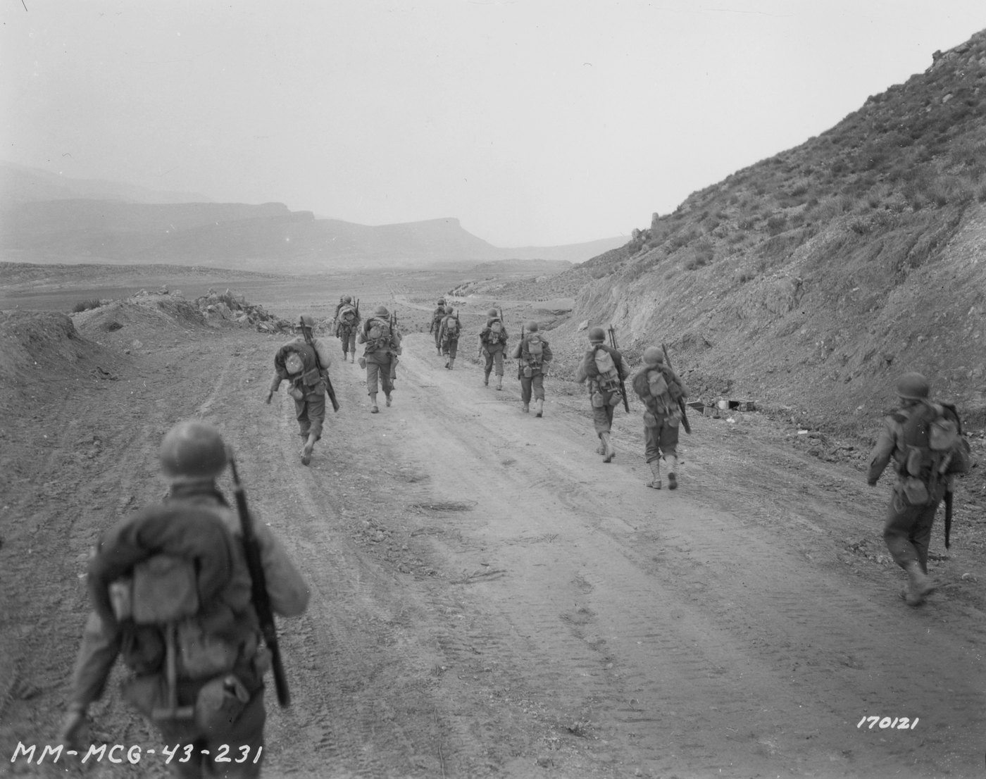 Black and white photograph: a line of American infantrymen with packs and rifles walks away from the camera along a dirt road through a wide, dry valley between hills.