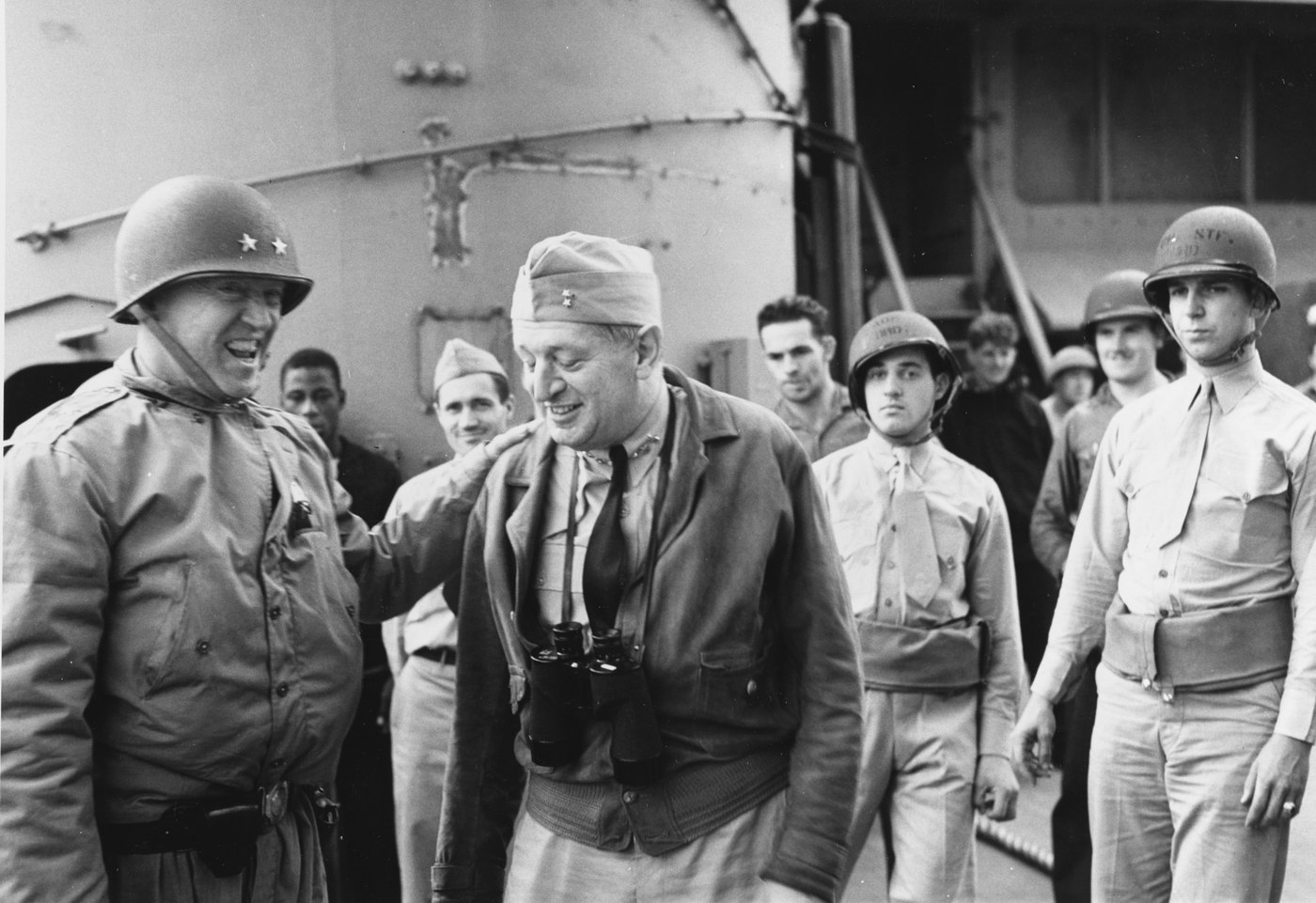 Black and white photograph on the deck of a warship: a laughing general in a steel helmet with two stars rests his hand on the shoulder of a smiling admiral wearing binoculars, with sailors and soldiers behind them.