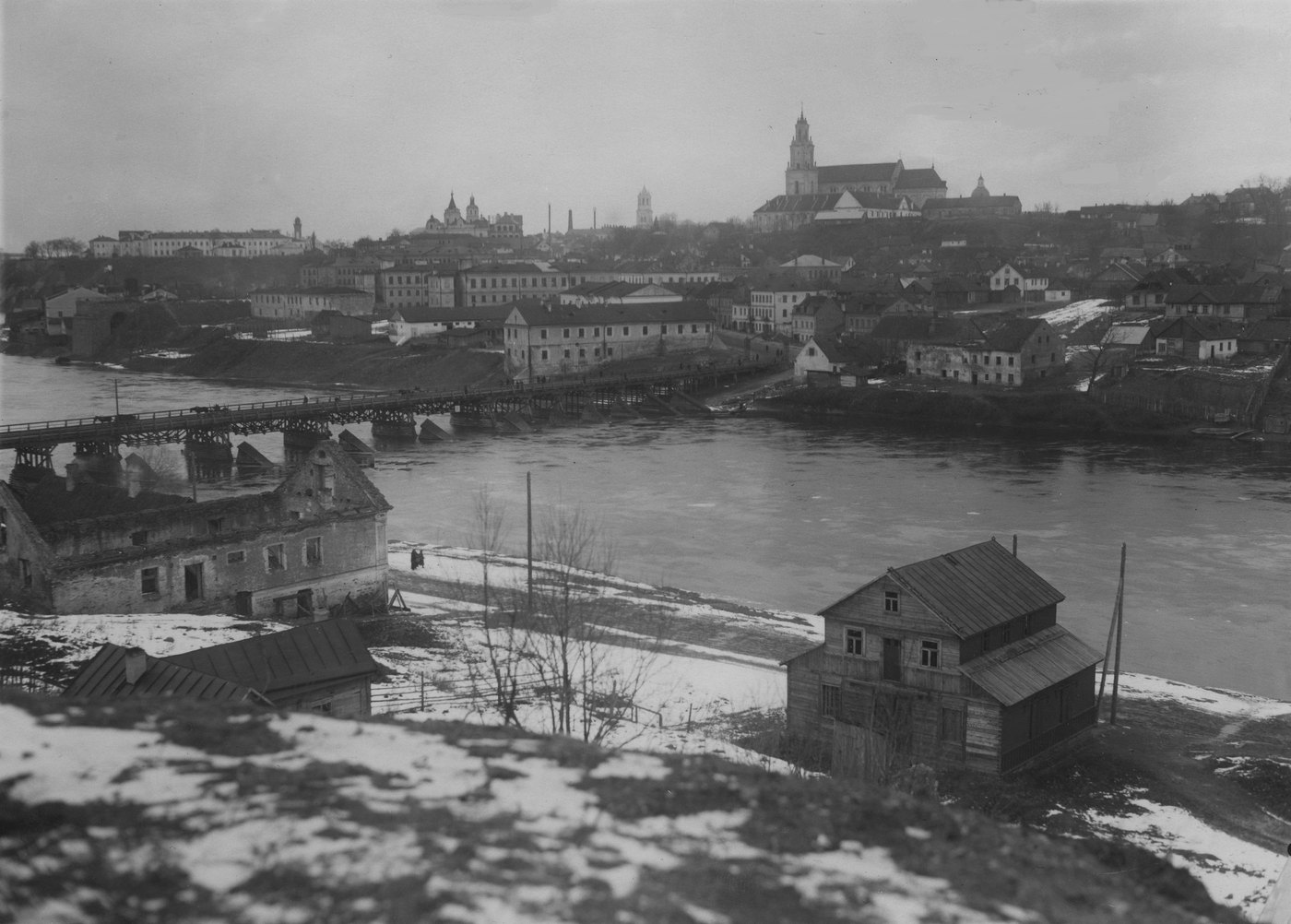Black and white view across a wide river to a town on a high bank with churches and towers, a long wooden bridge beside the stumps of older piers, and snow on the near bank