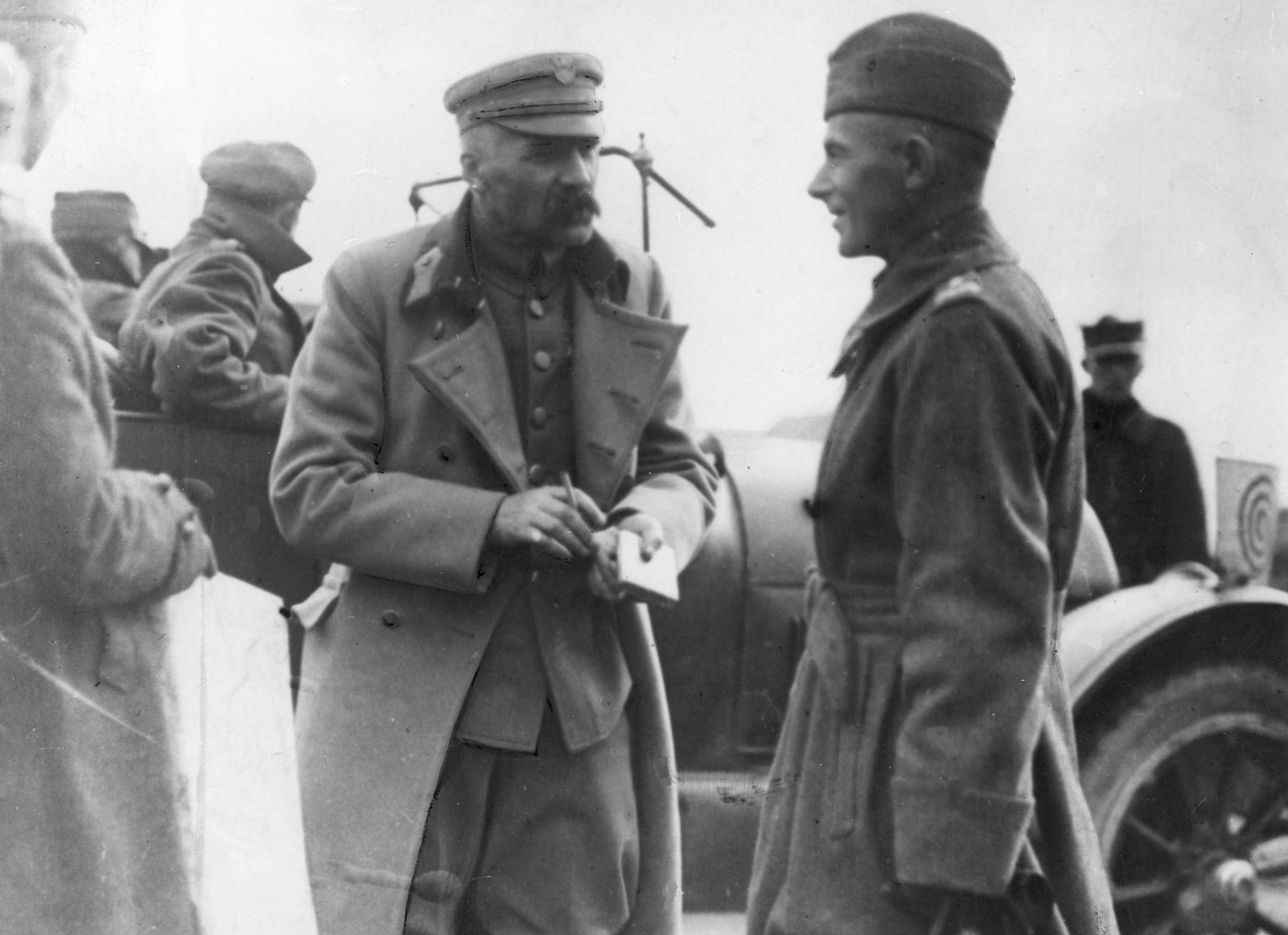 Black and white photograph of a moustached man in a peaked cap and greatcoat holding a pencil and notebook, talking to a smiling younger officer in a field cap beside a car