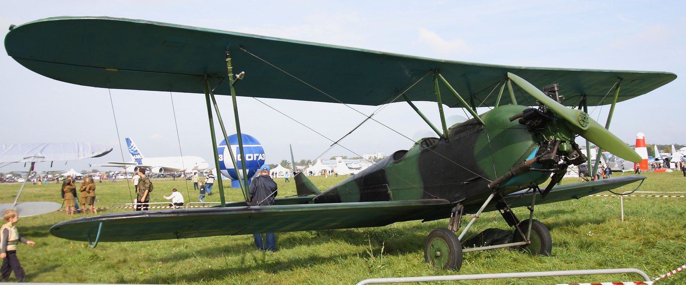 A green and black camouflaged biplane with two open cockpits and a radial engine standing on grass, with visitors and modern airliners in the background
