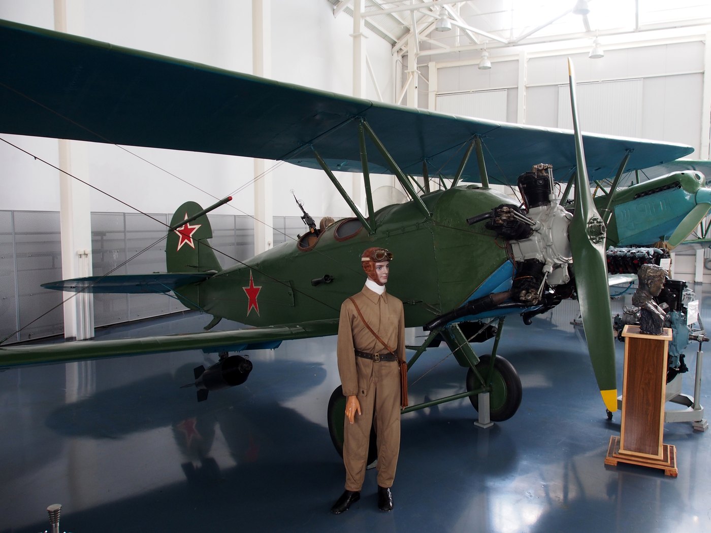 Green Po-2 biplane with red stars seen from the front quarter, its radial engine and two-blade propeller exposed, a bomb under the lower wing and a mannequin in a leather flying helmet and overalls standing in front