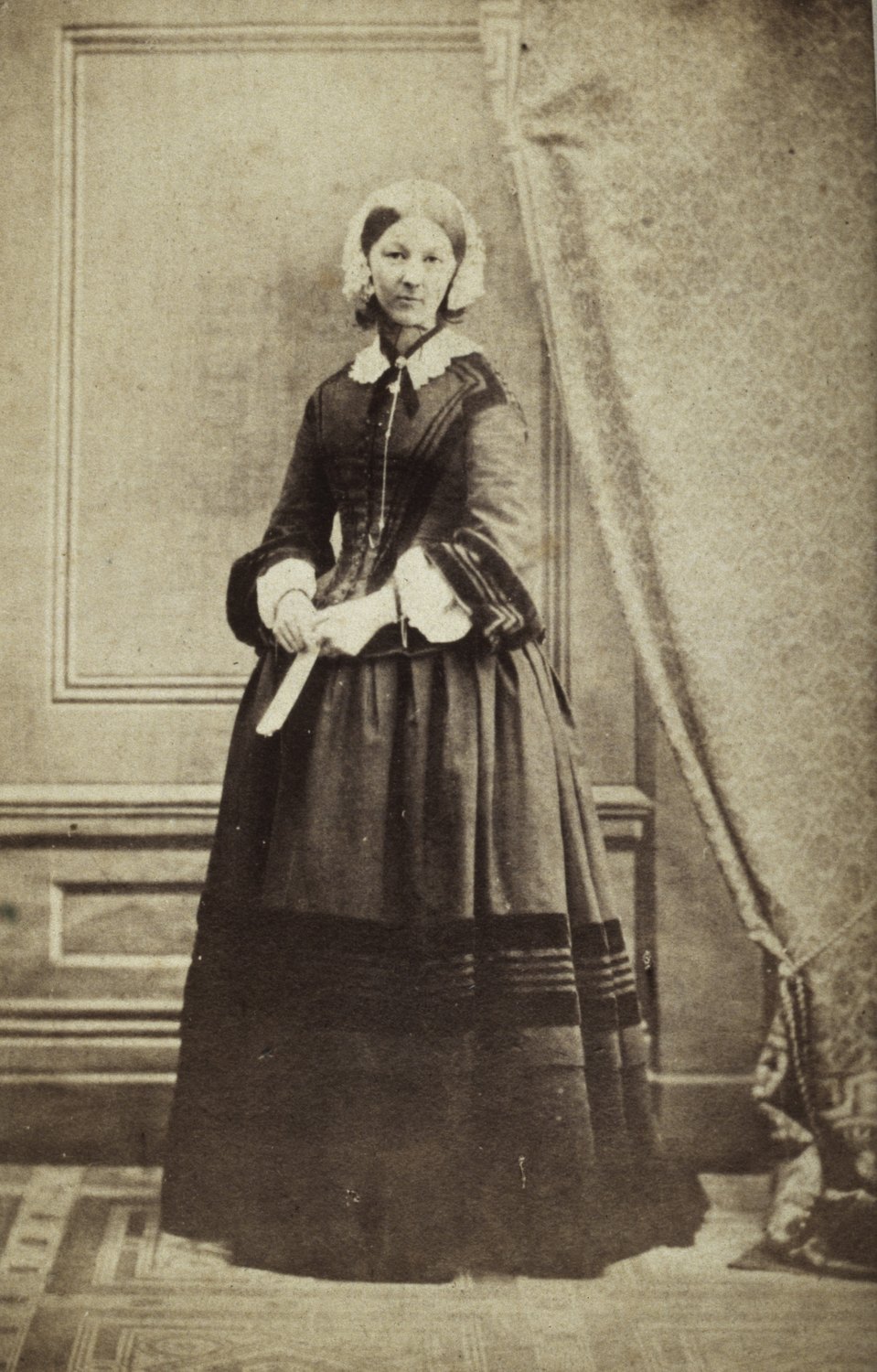 Full-length sepia photograph of a woman in a dark dress with a lace collar and white cap, standing in front of a paneled wall and a curtain