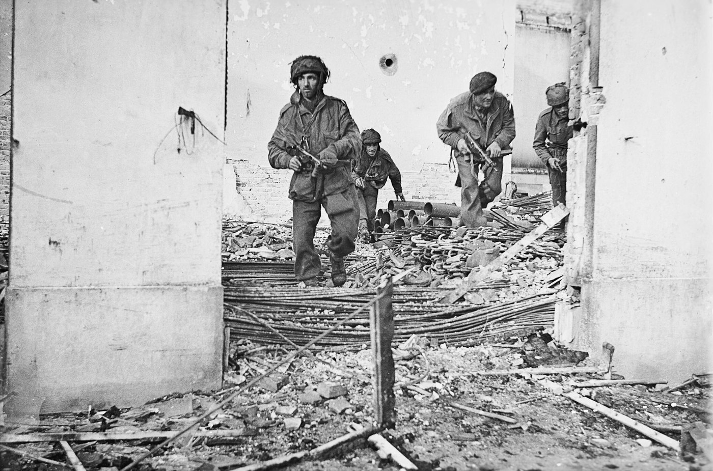 Four British paratroopers with Sten guns and rifles move through the rubble of a shell-damaged house, the leading man in a netted helmet, another in a beret.