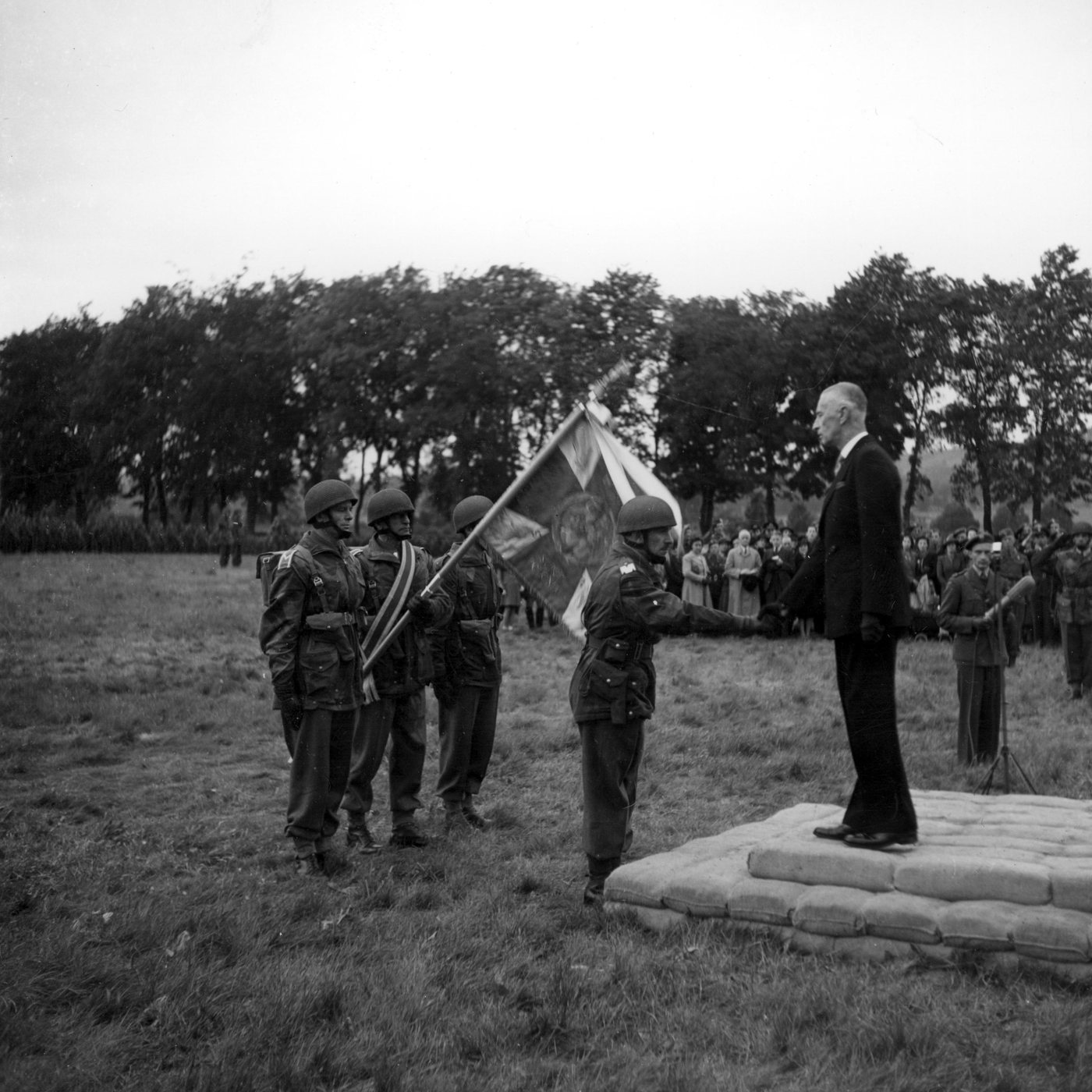 A man in a dark suit on a low platform faces paratroopers holding a standard on a parade ground
