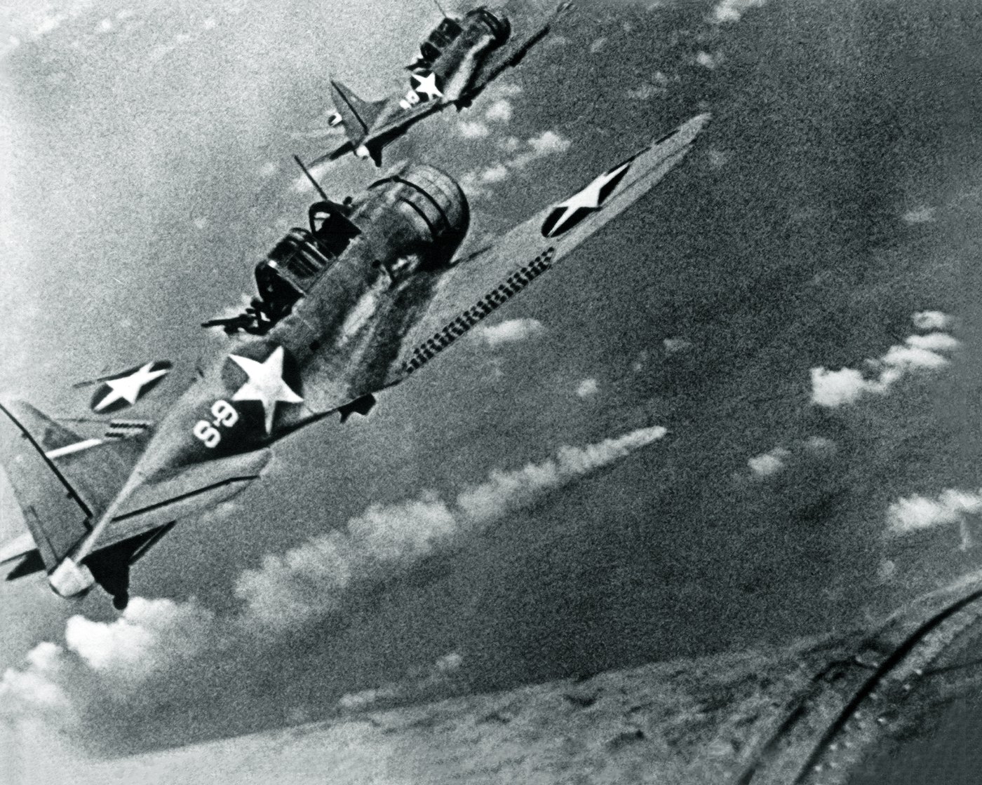 Black and white photograph from the air: two U.S. Navy dive bombers with white star insignia fly over the sea toward a burning warship far below.