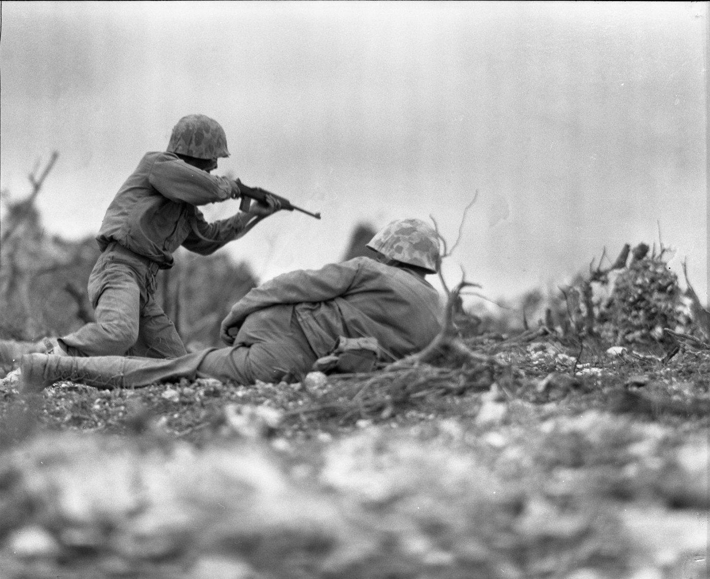 Black and white photograph: a U.S. Marine in a camouflage helmet fires a submachine gun over another Marine lying on rocky, shell-torn ground.