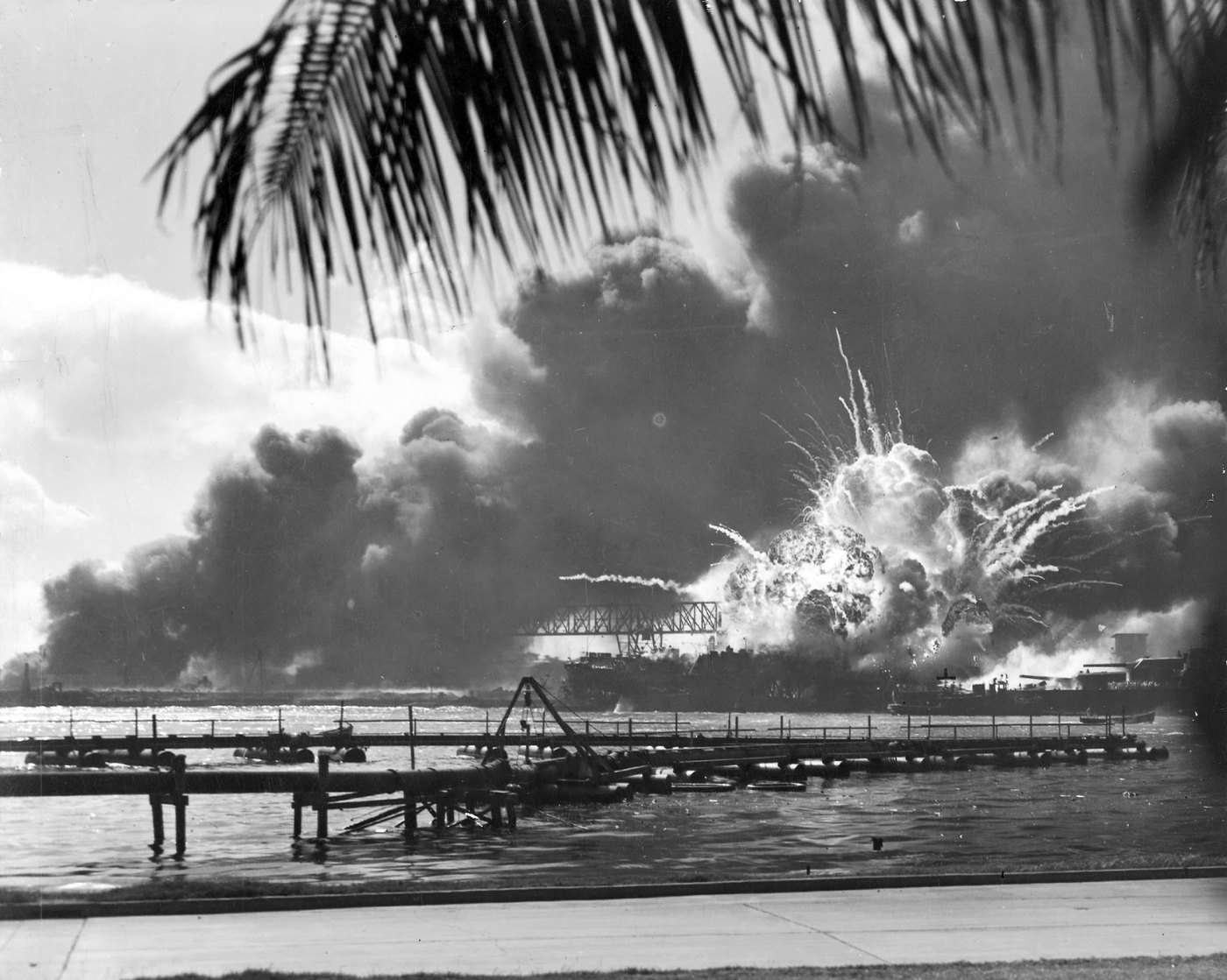 Black and white photograph: a huge fireball and black smoke rise from a destroyer in a floating dry dock, seen across the water past palm fronds.