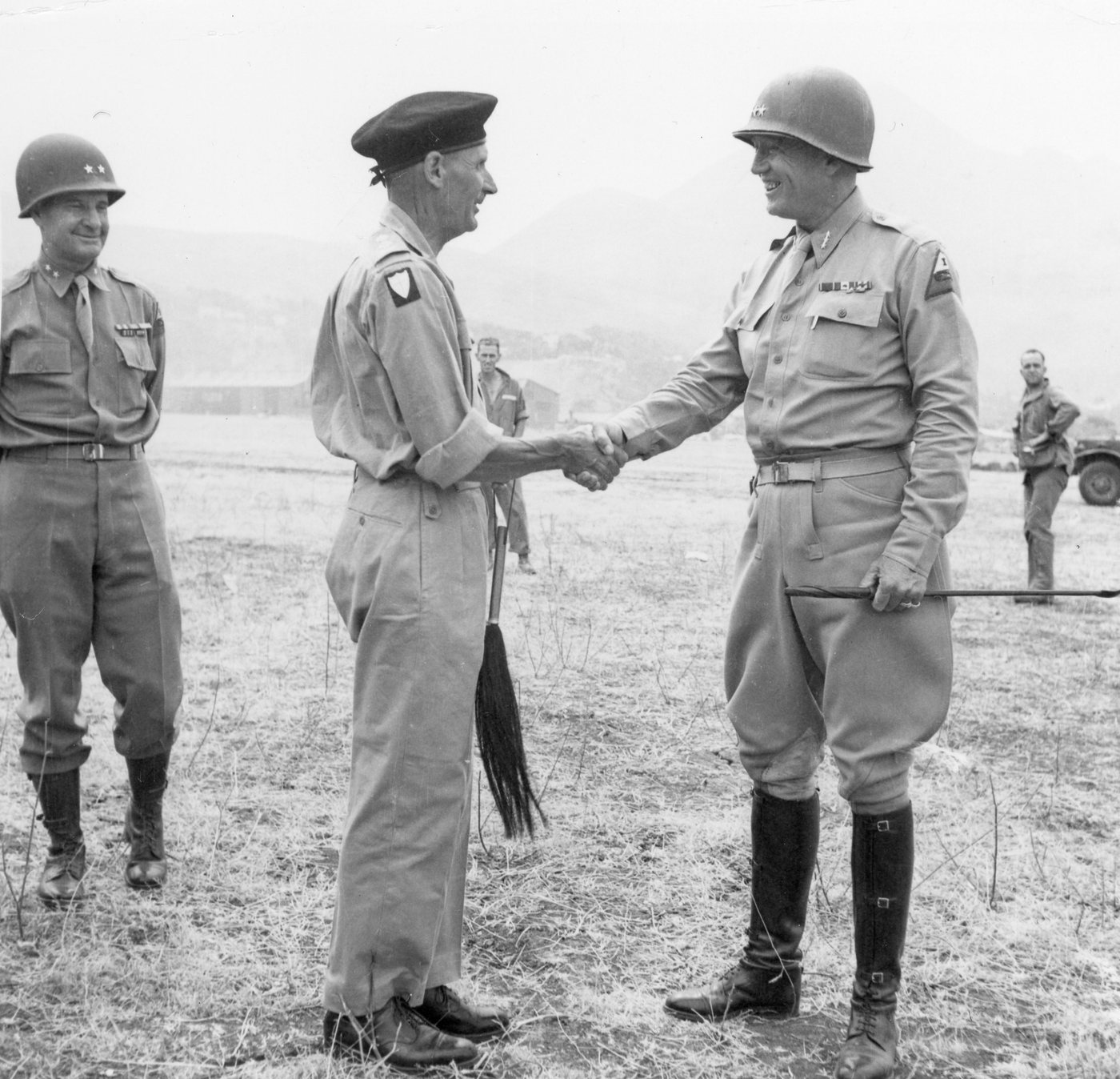 Montgomery in a beret and Patton in a helmet shaking hands on an airfield, with another officer behind them