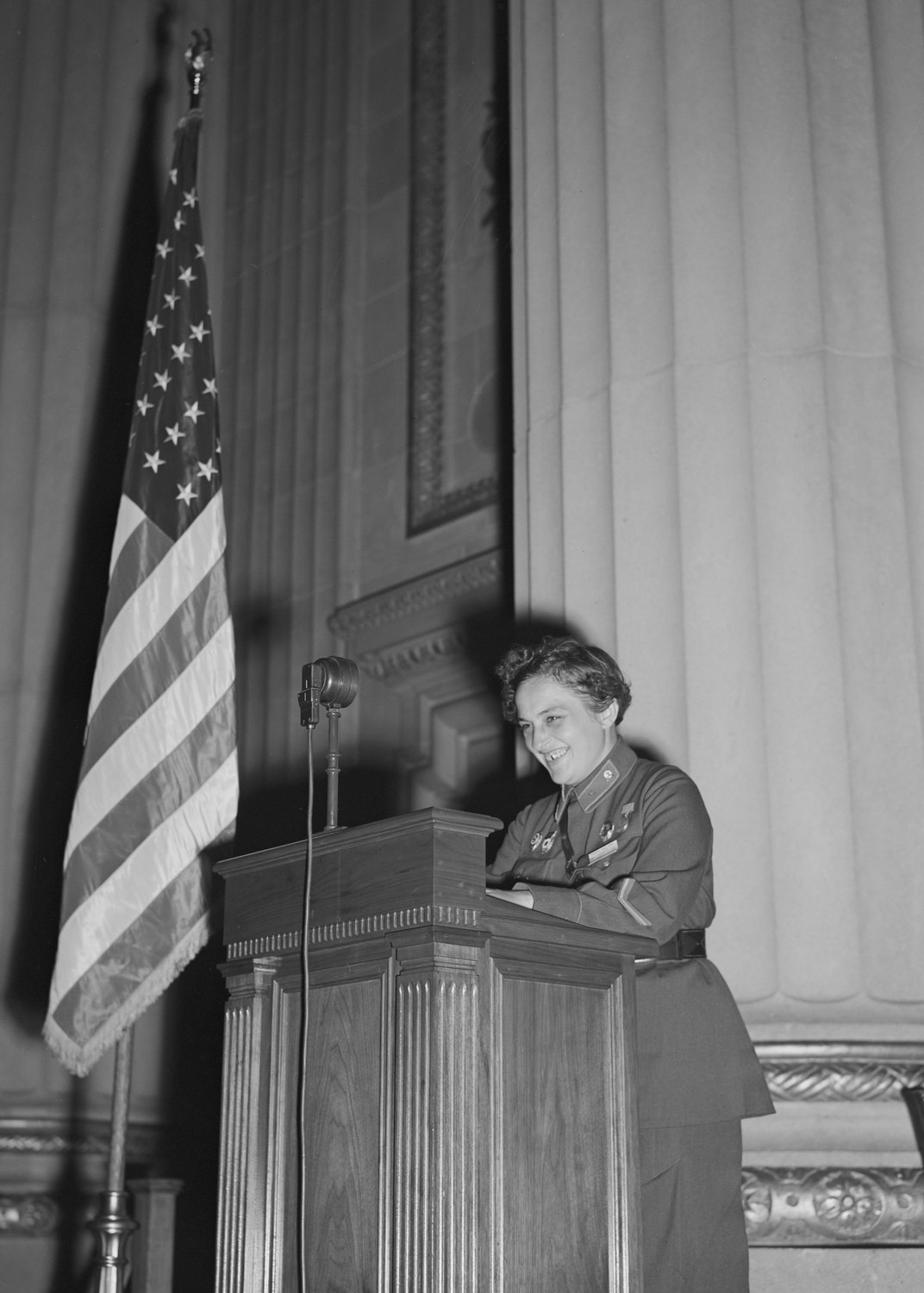 A smiling young woman in Soviet army uniform speaks at a wooden lectern with a microphone, an American flag beside her and tall columns behind