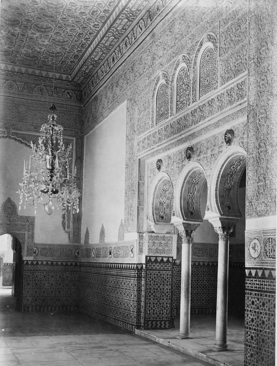 Old black and white photograph of a palace hall with carved stucco walls, horseshoe arches on slim columns, tiled dados and a chandelier.