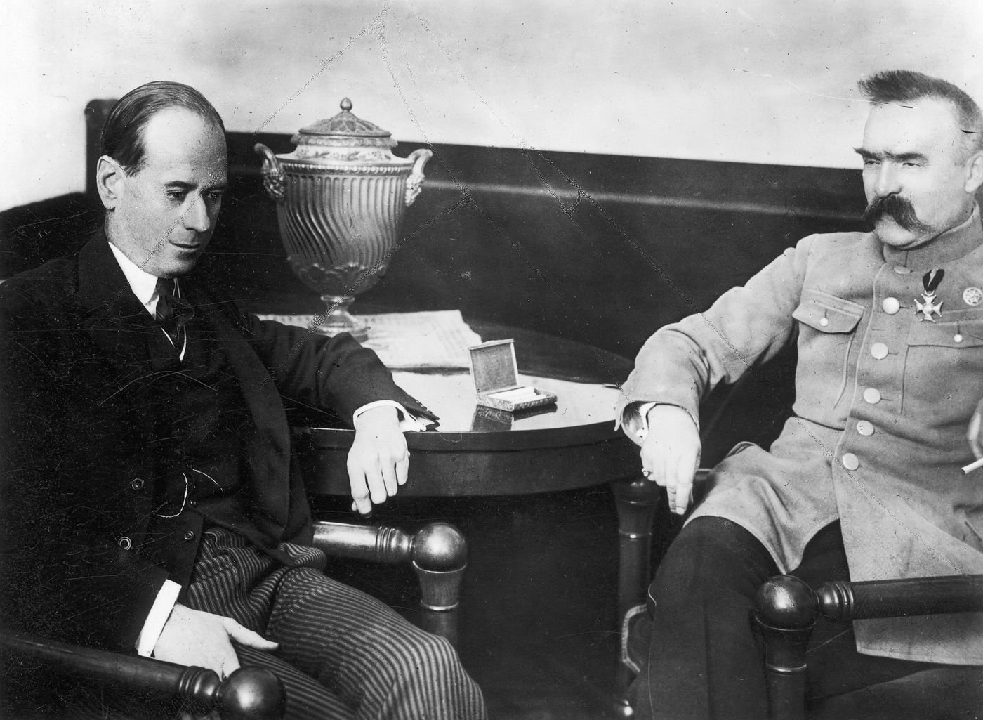 Black and white photograph of Józef Piłsudski in a uniform jacket with a cross on his chest, seated at a round table beside a man in a dark suit