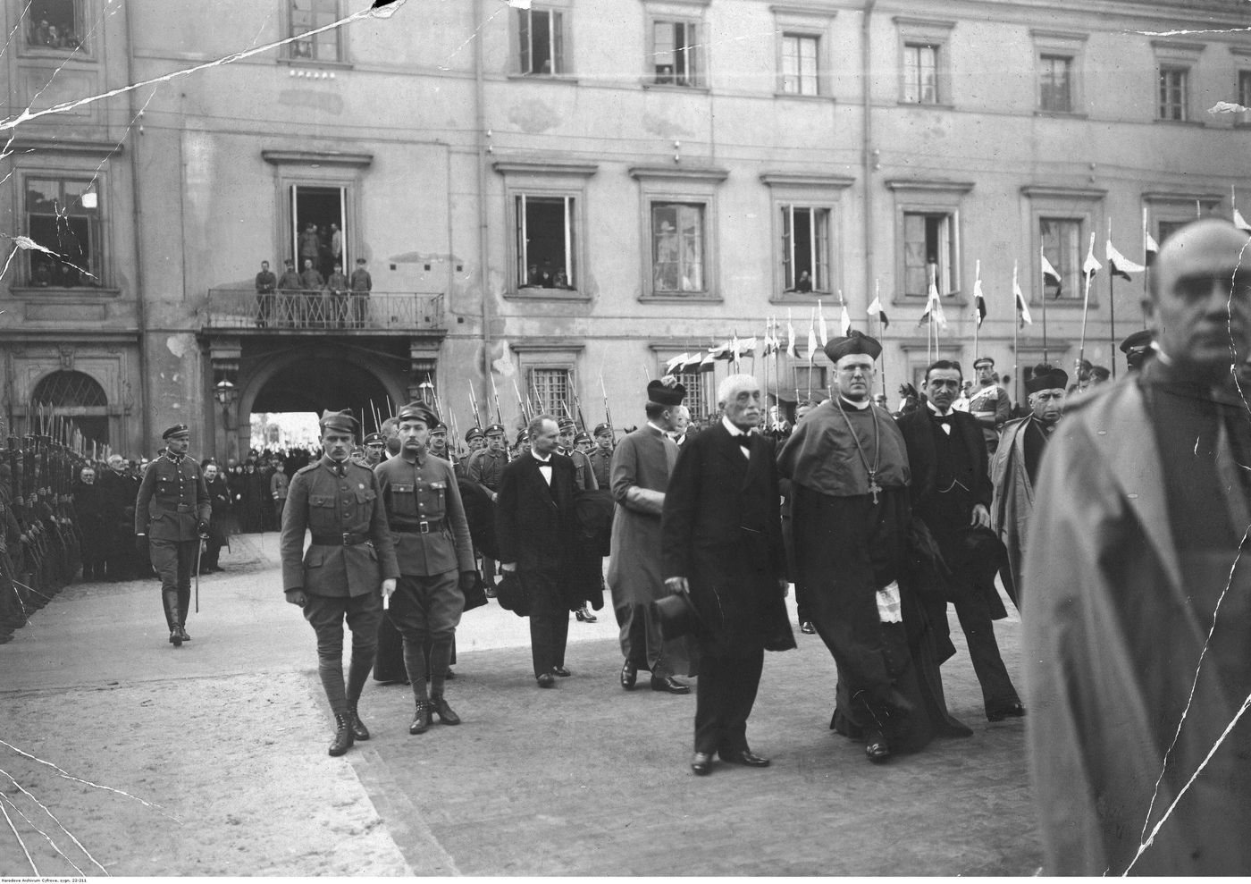 Black and white photograph of men in dark coats and a bishop in robes walking past lines of soldiers in a palace courtyard, with flags behind them