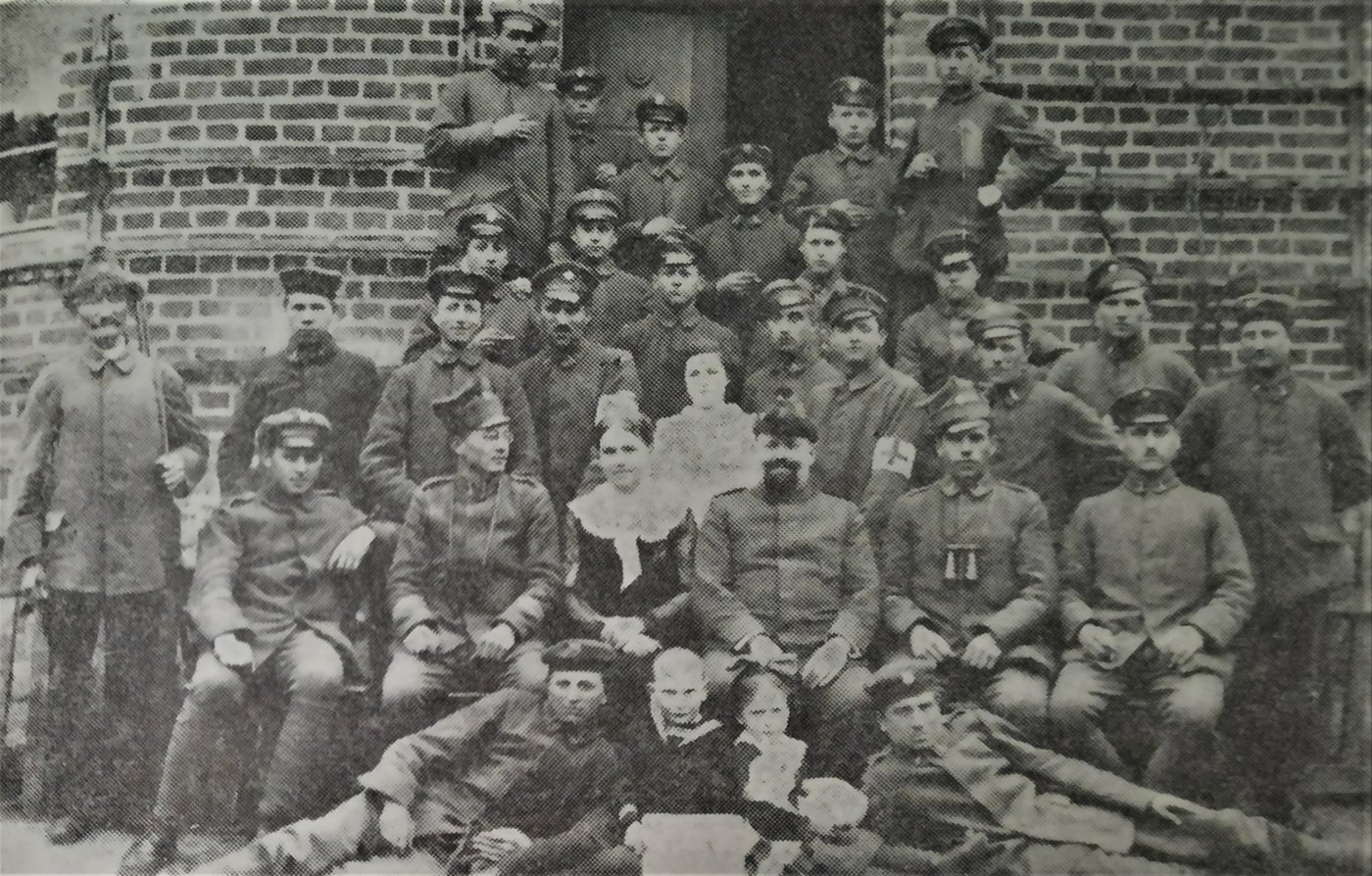 Old group photograph of about thirty men in military caps and tunics, a few with armbands and binoculars, with two women and small children, posed in front of a brick building
