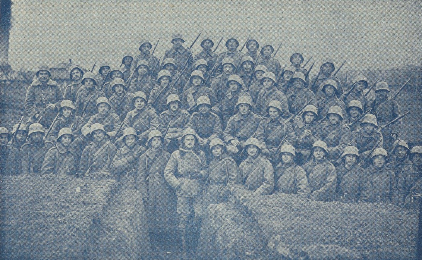 Group photograph of several dozen soldiers in steel helmets with rifles and fixed bayonets, standing and kneeling in and around a trench, with an officer in a fur collared coat in the center