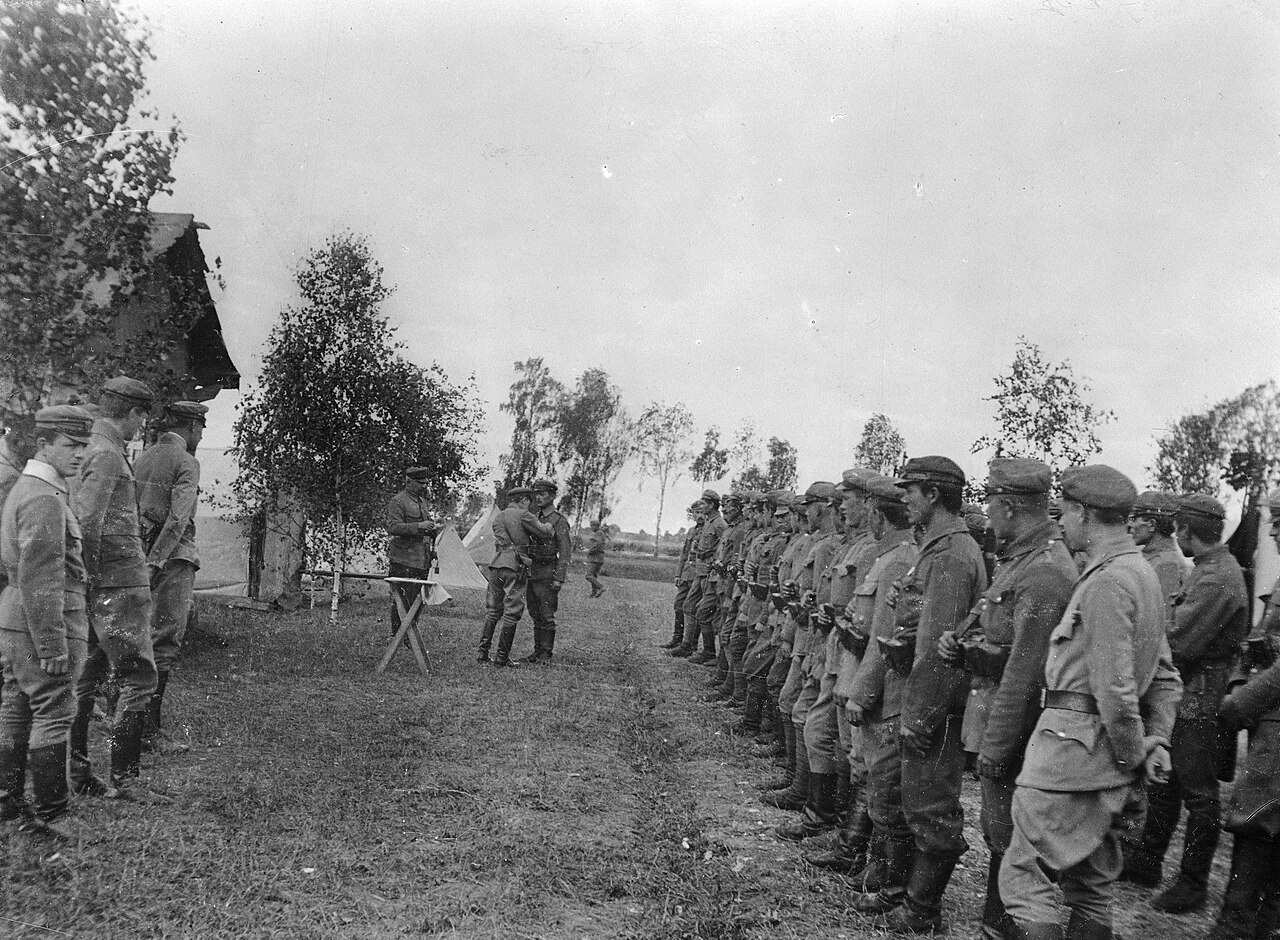 Black and white photograph of a decoration ceremony in a field: officers by a small table, one pinning a decoration on a soldier, between two long lines of Legion soldiers in caps