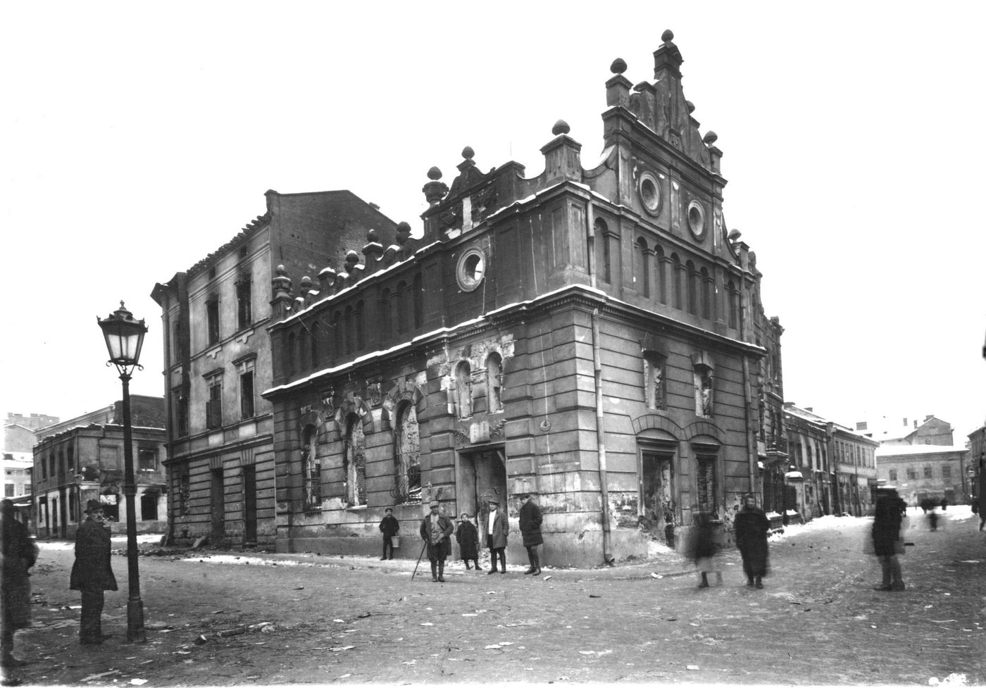 Czarno-białe zdjęcie wypalonej synagogi z ozdobną fasadą zwieńczoną szczytem, pustymi, osmalonymi otworami okiennymi i poplamionymi kamiennymi murami; przed nią kilku mężczyzn i chłopców na zaśnieżonej ulicy, po lewej latarnia