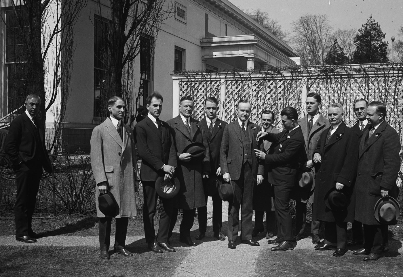 A group of men in dark suits and overcoats, most holding hats, stand on a garden path in front of a pale building and a lattice fence while one man pins a small flower to the lapel of the man in the center