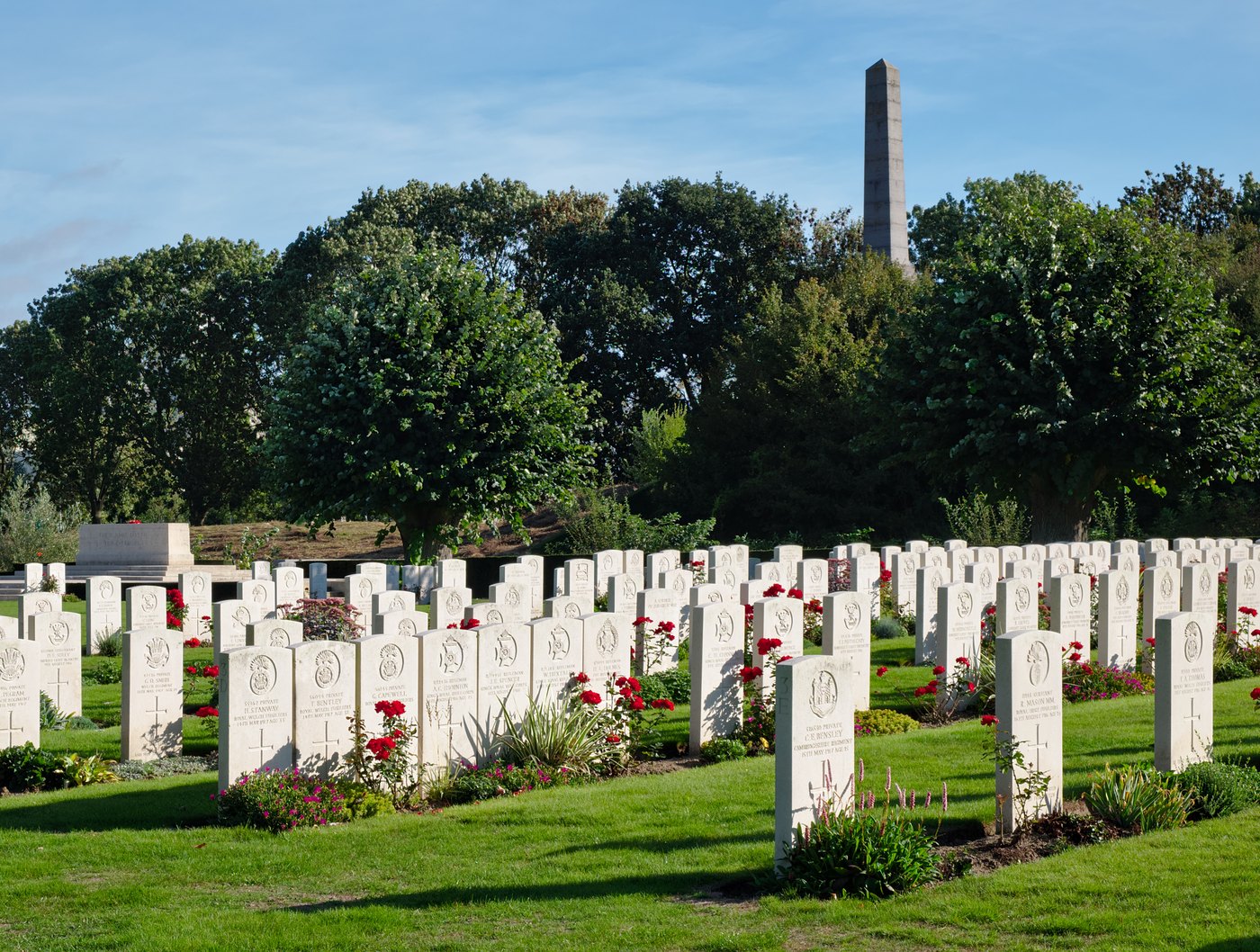 Rows of white headstones on a mown lawn, with red flowers planted between them, trees behind and a tall stone obelisk rising above the trees under a blue sky