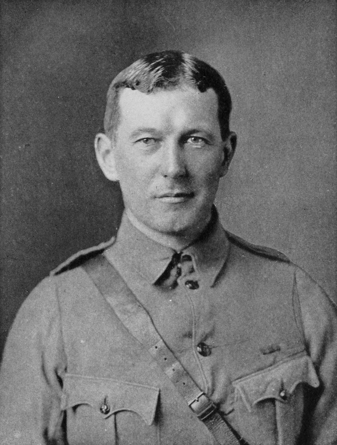 Black and white head and shoulders portrait of a clean shaven officer with short parted hair, in a buttoned service tunic with a leather cross strap