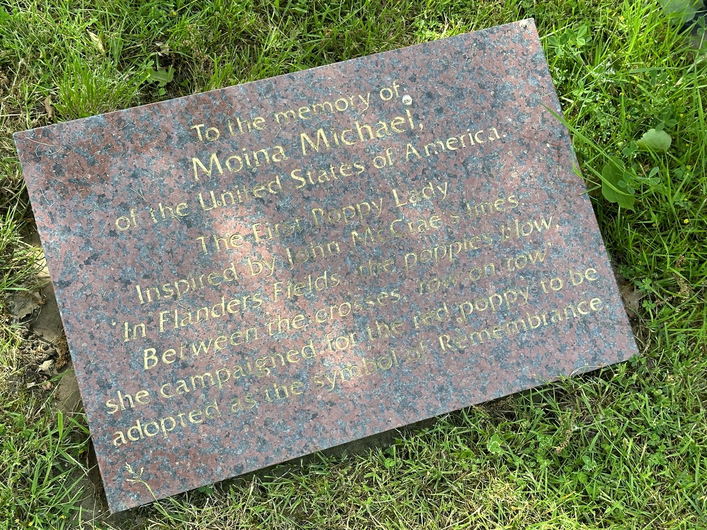 A speckled red granite plaque lying in grass, engraved in gold letters to the memory of Moina Michael of the United States of America, the first Poppy Lady