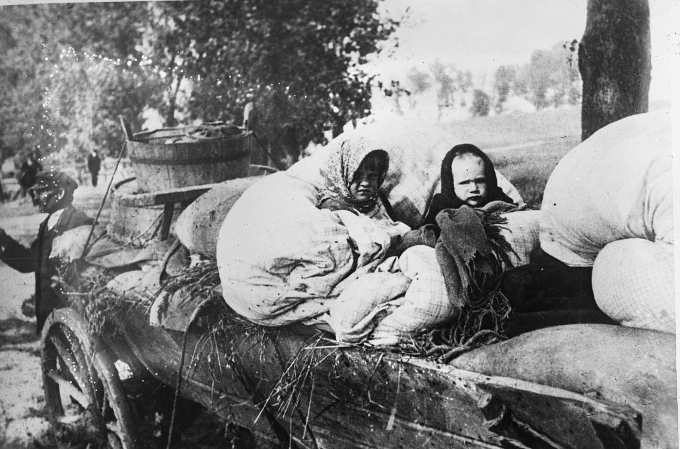 Czarno-biała fotografia dwojga małych dzieci w chustkach, siedzących wśród tobołków i worków ułożonych na drewnianym wozie.