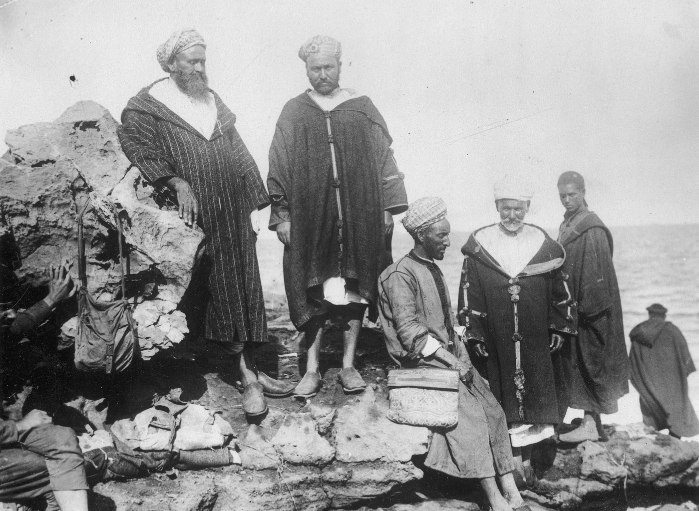 Black and white photograph of five men in hooded robes and turbans standing and sitting on rocks by the sea.