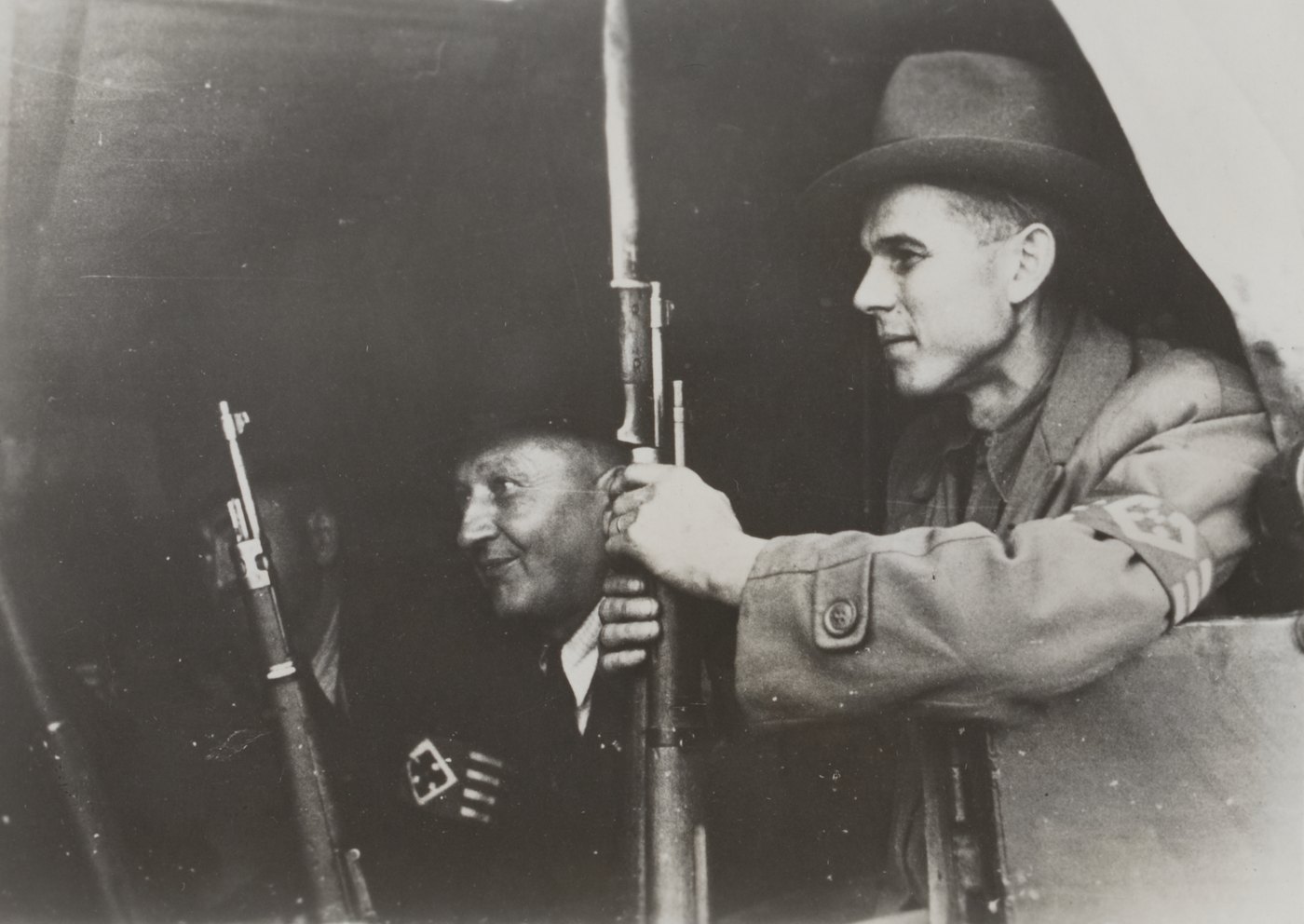 Black and white photograph of two armed civilians in a truck, a younger man in a felt hat and a jacket with an armband holding a rifle with a fixed bayonet, an older man beside him, another rifle in the foreground