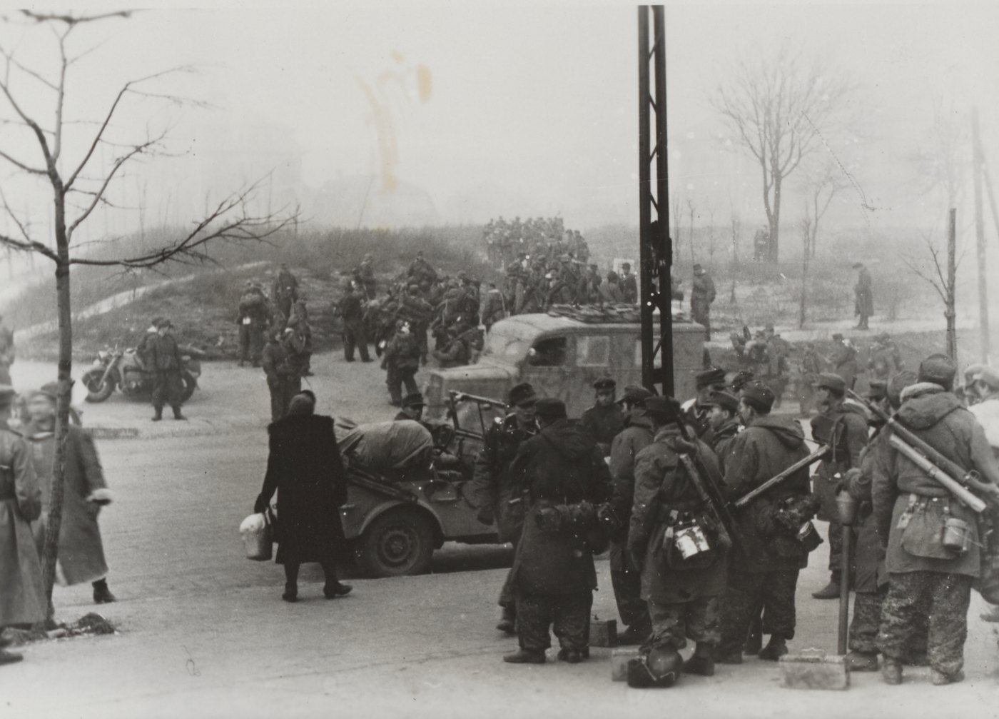 Black and white photograph of German soldiers in winter parkas, field caps and camouflage trousers, carrying submachine guns and antitank rocket launchers, gathered at a street corner in fog beside a car loaded with kit, a woman in a dark coat with a bag crossing the road and a column of troops on a slope behind