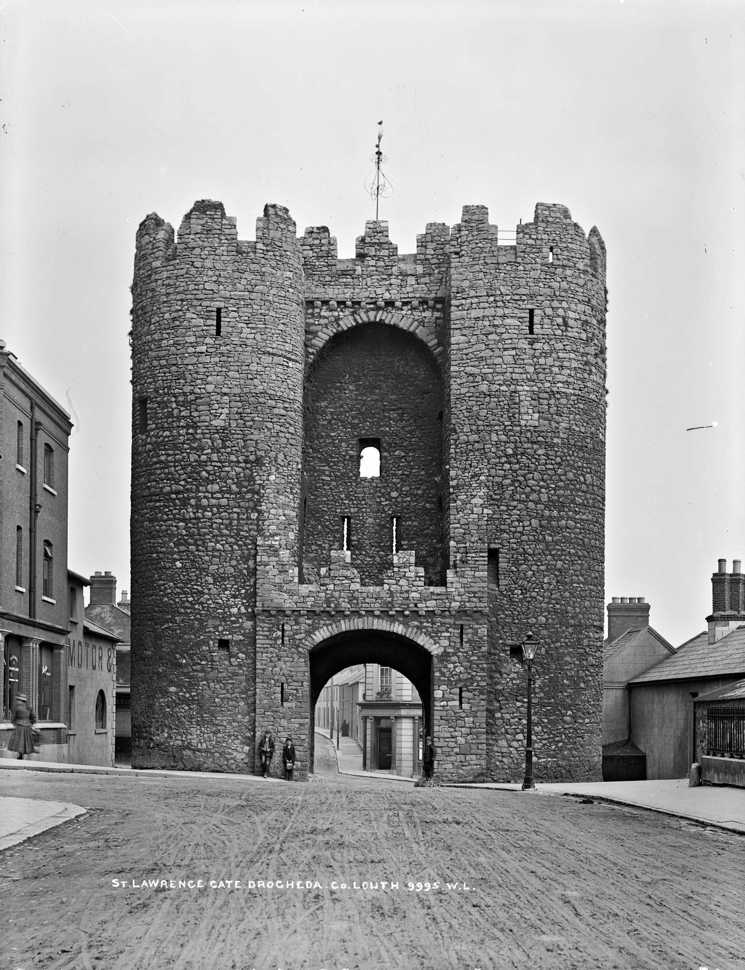 Black and white photograph of a tall medieval gate building with two round towers joined by a high arch and battlements, a smaller arched gateway at street level, a few people standing beneath it and town houses on either side of an unpaved street
