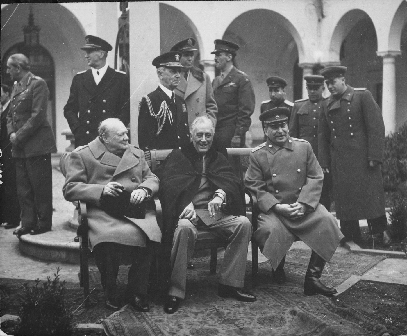 Czarno-białe zdjęcie trzech przywódców siedzących obok siebie na patio, Churchilla w płaszczu, Roosevelta w ciemnej pelerynie i Stalina w mundurze marszałka, a za nimi pod kamiennymi arkadami stoją oficerowie brytyjscy, amerykańscy i radzieccy.