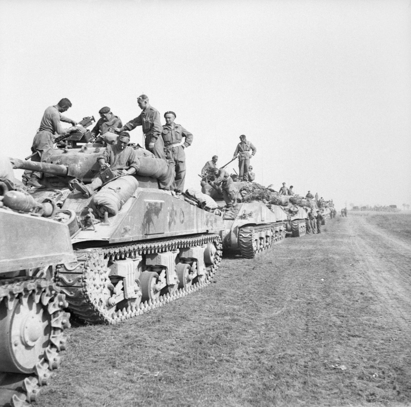 A column of Sherman tanks parked along a road with crews sitting on the turrets