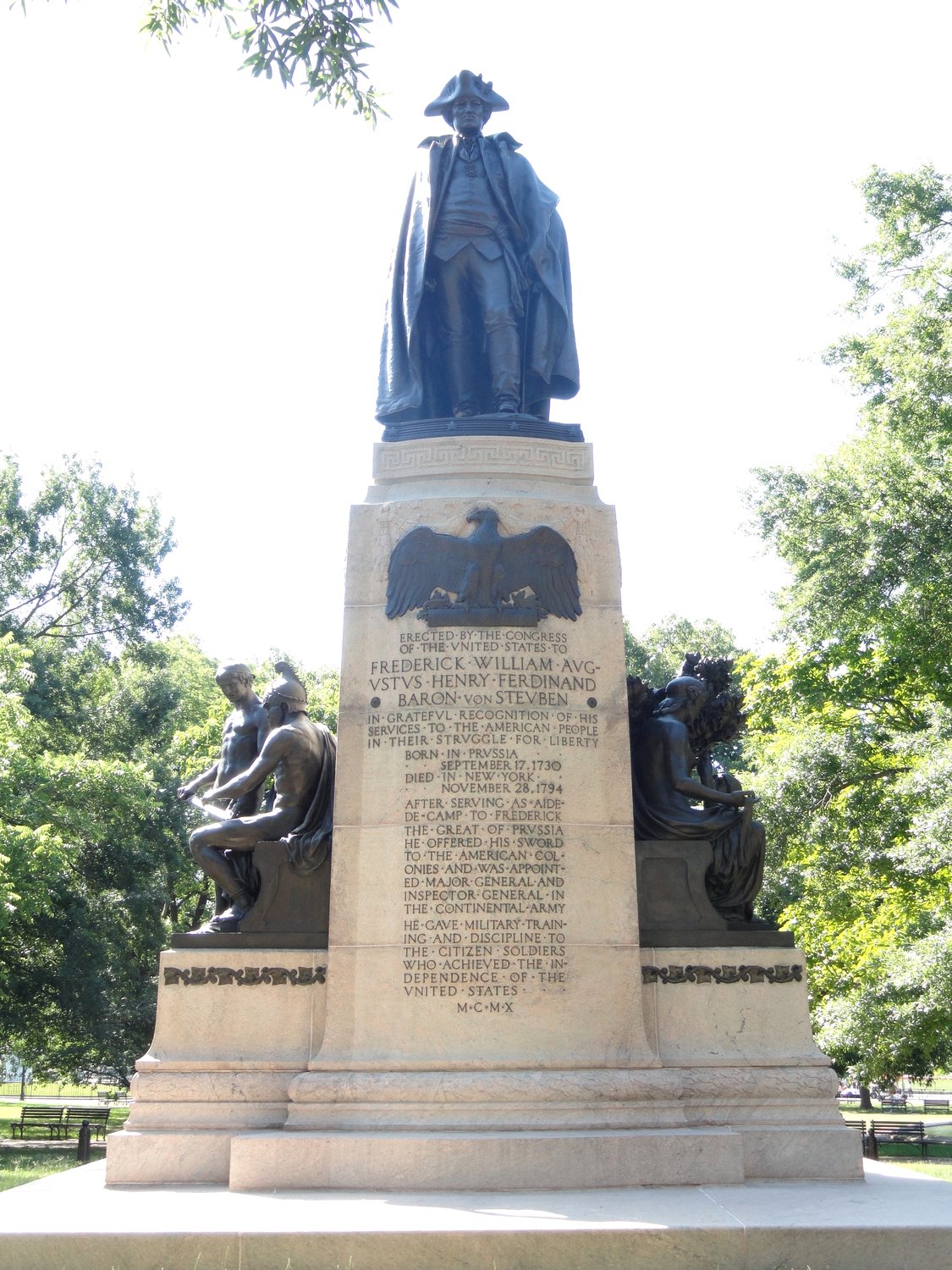 Bronze statue of Steuben in a long cloak and tricorne hat on a tall granite pedestal among trees
