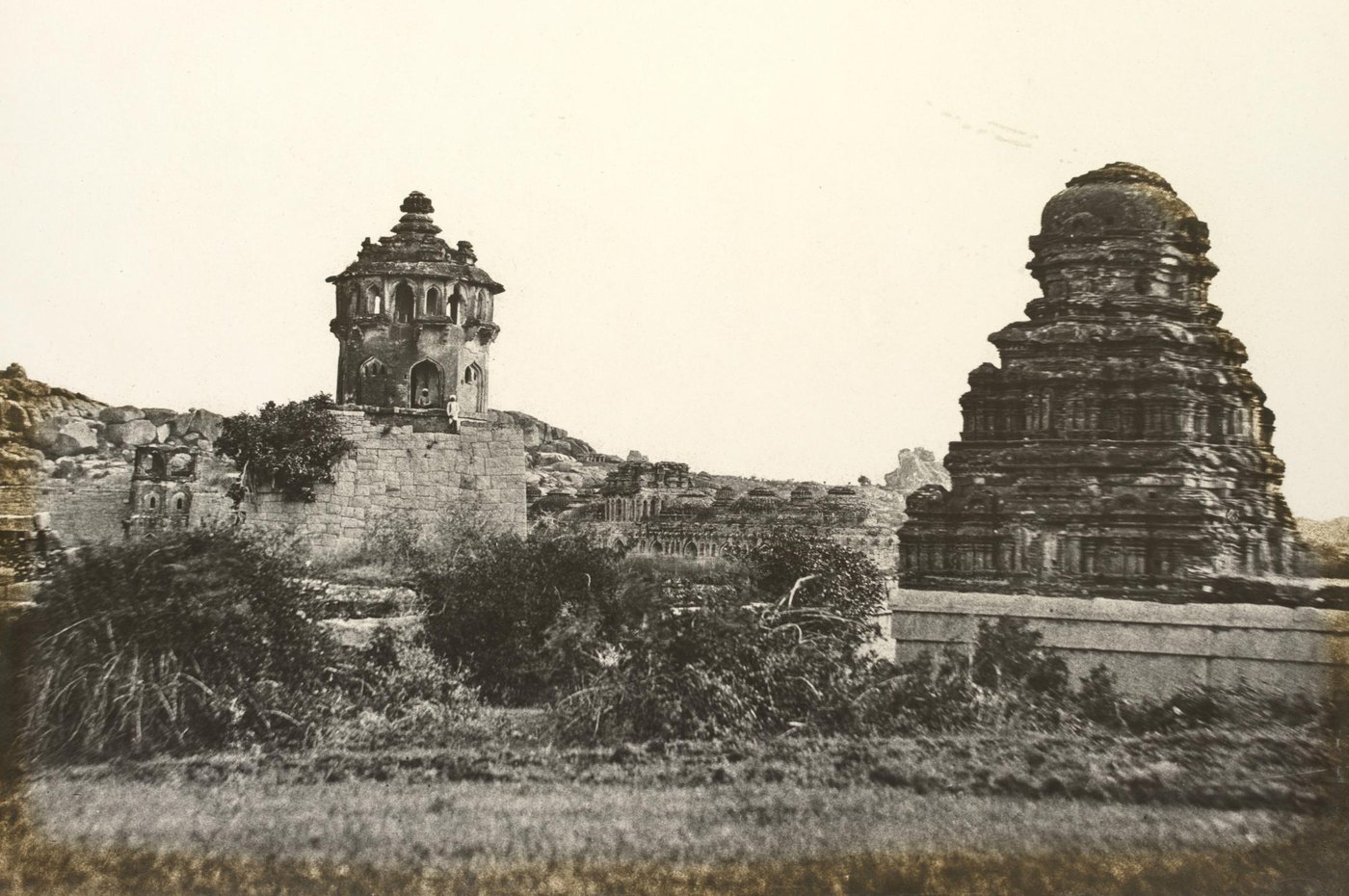 Old sepia photograph of ruins: an octagonal domed watchtower on a stone wall at left, a long domed building behind, and a weathered temple tower at right above scrub