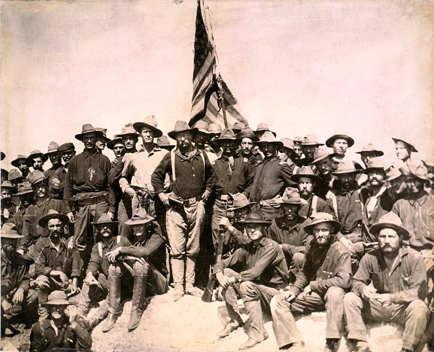Group photograph of Rough Riders gathered on a hilltop under an American flag, Roosevelt at the centre