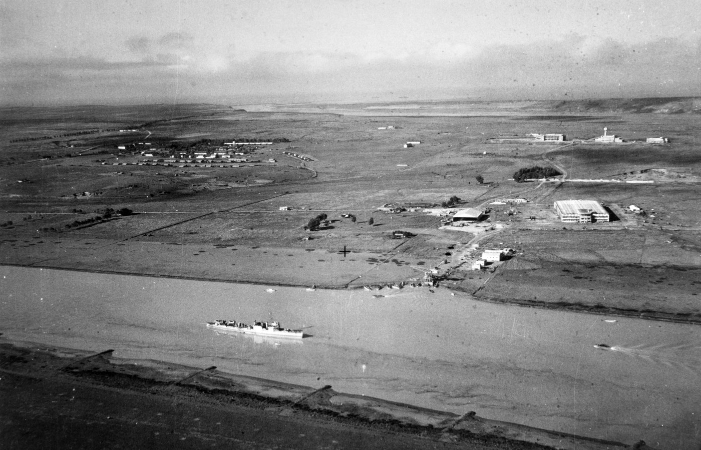 Aerial photograph of a small warship anchored in a wide river, with landing craft beached on the far bank in front of an airfield with hangars, aircraft and barracks on a flat plain