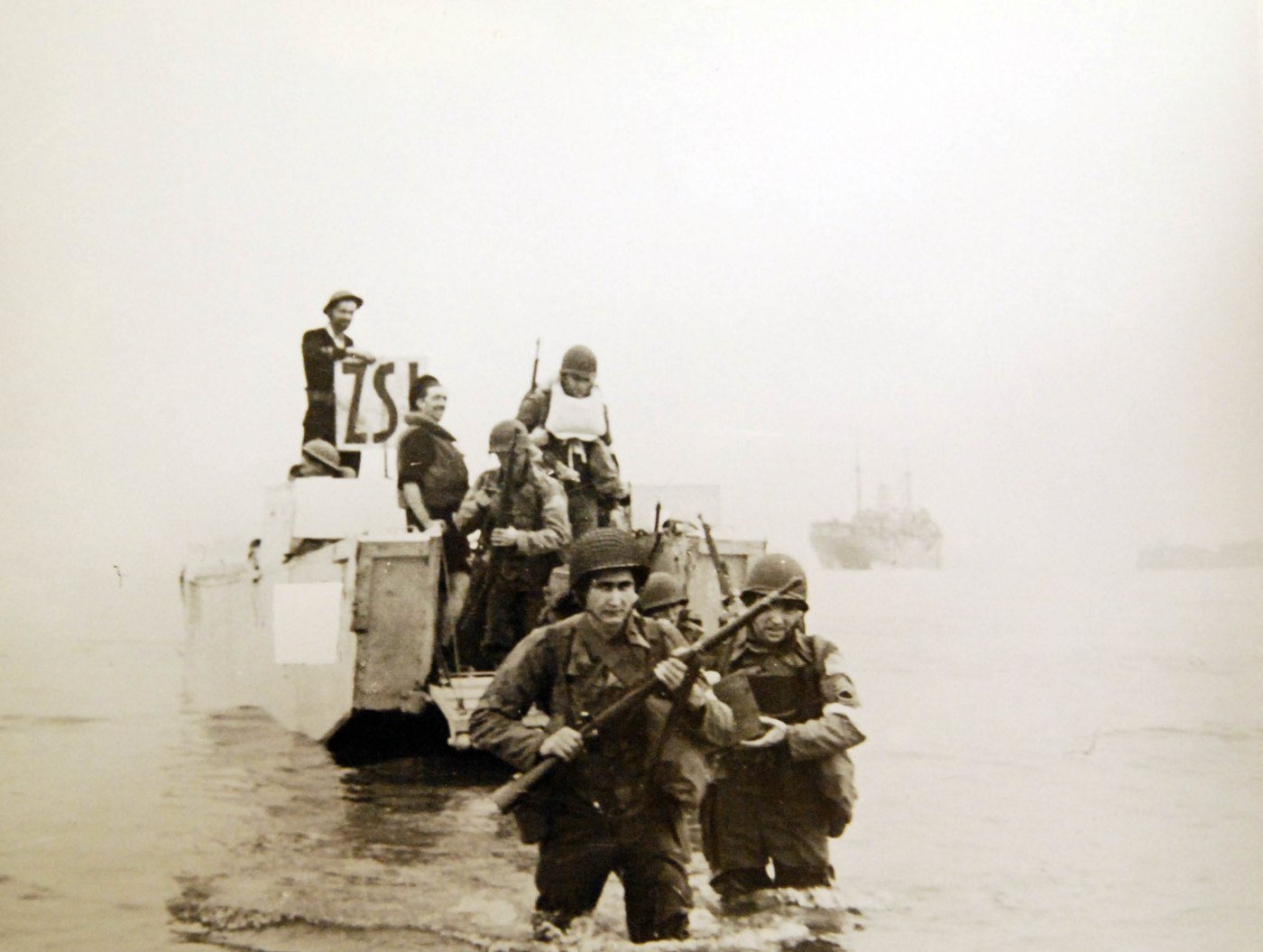 Two American soldiers in helmets wade through shallow water towards the camera carrying rifles, as more soldiers climb down from a landing craft behind them and a transport ship stands in the haze