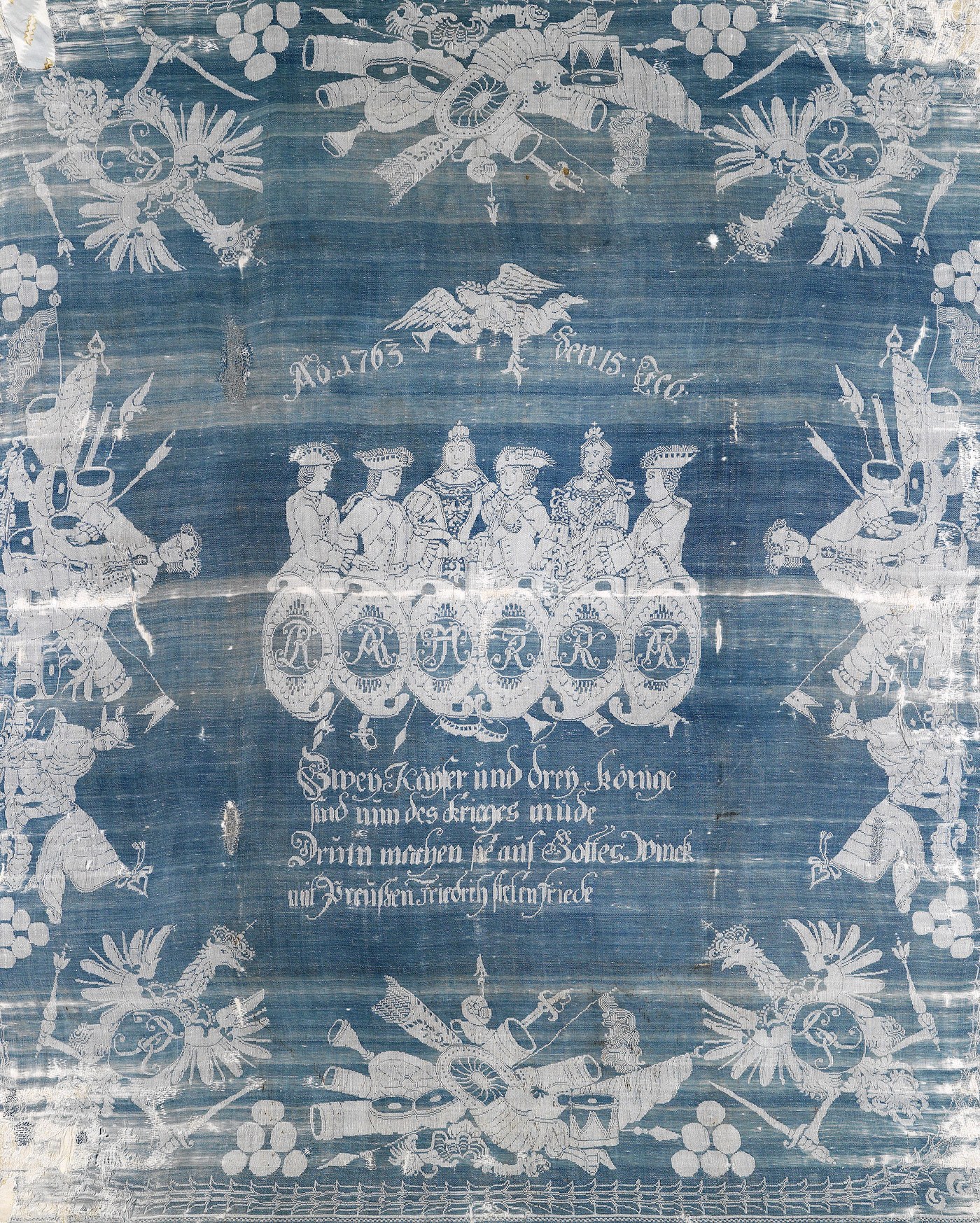 Blue and white woven linen cloth: in the center six crowned or hatted figures stand above six oval monograms, with the date 15 February 1763 and a German verse below, framed by a Prussian eagle and trophies of flags, drums and cannon.