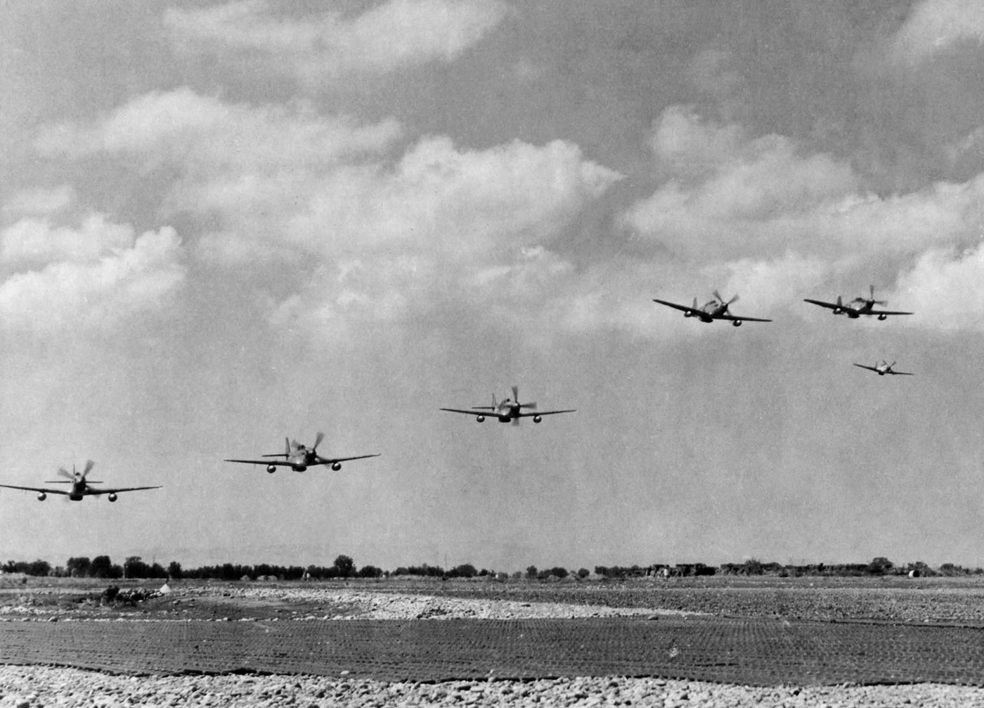 Black and white photograph of P-51 Mustang fighters with drop tanks taking off from a dusty airfield